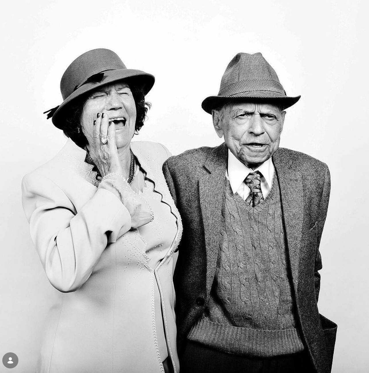 Black and white portrait of a laughing elderly woman in a brimmed hat beside a smiling elderly man in a fedora, tweed jacket and tie.