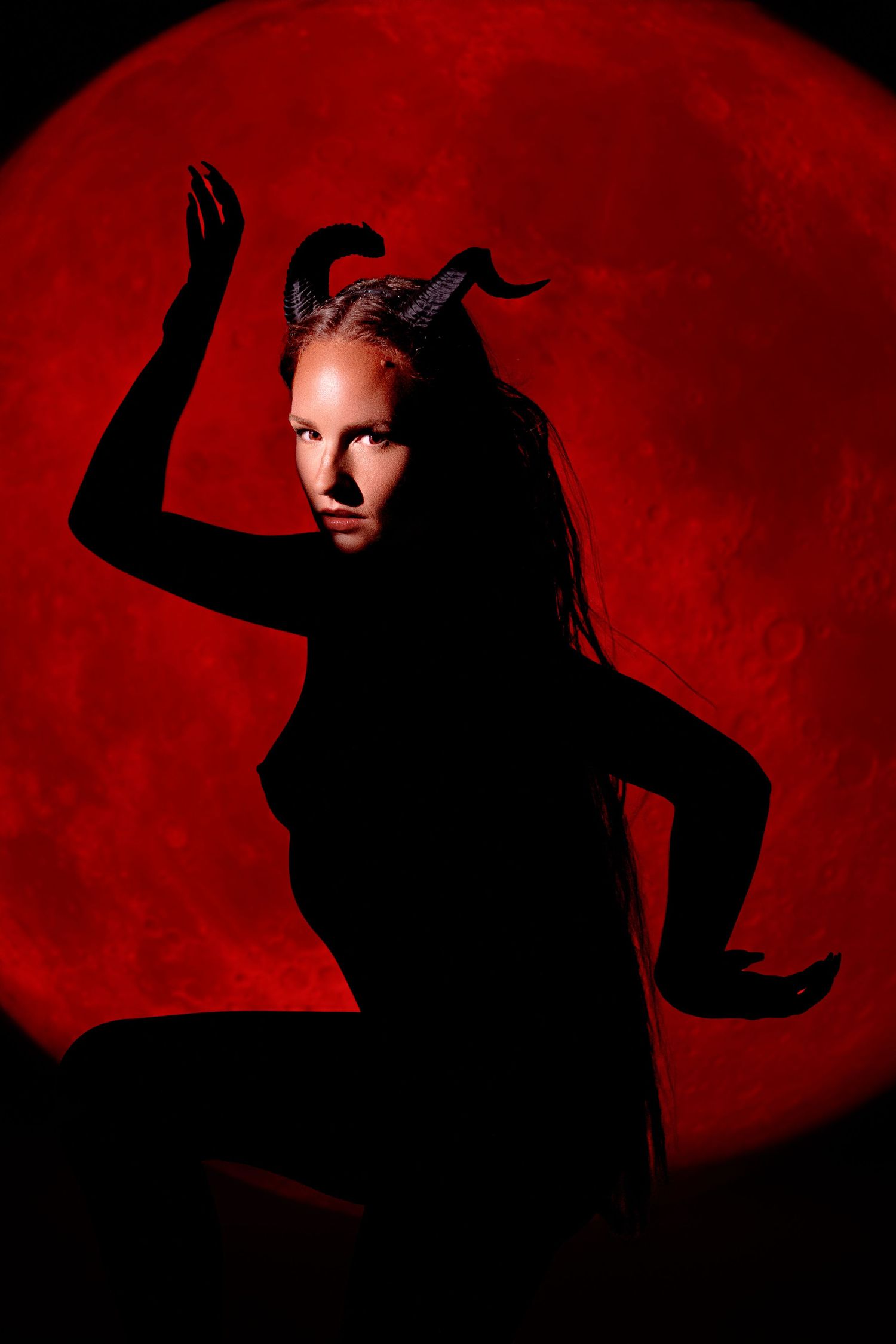A dramatic silhouette of a woman with devil horns posing against a glowing red full moon background.