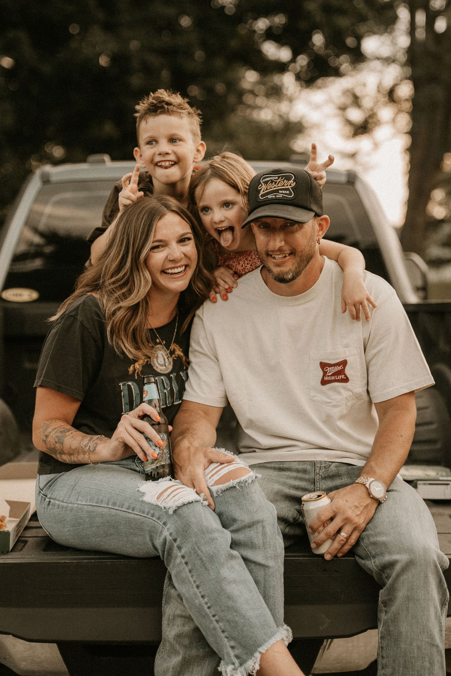 Happy family of four sitting on truck tailgate, parents laughing with two playful kids making funny faces outdoors.