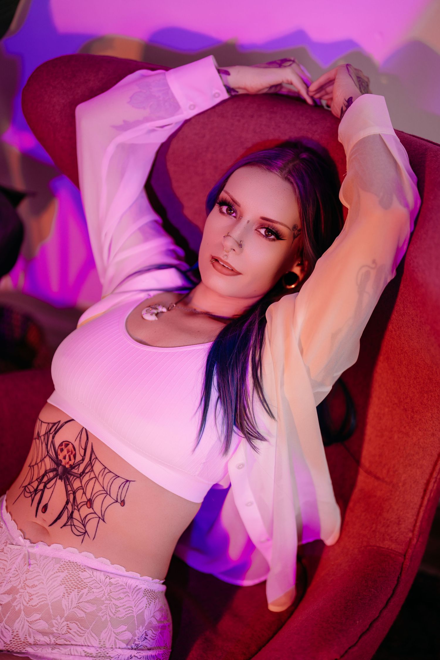 Person in white top with visible tattoo poses against red backdrop with purple lighting effects.