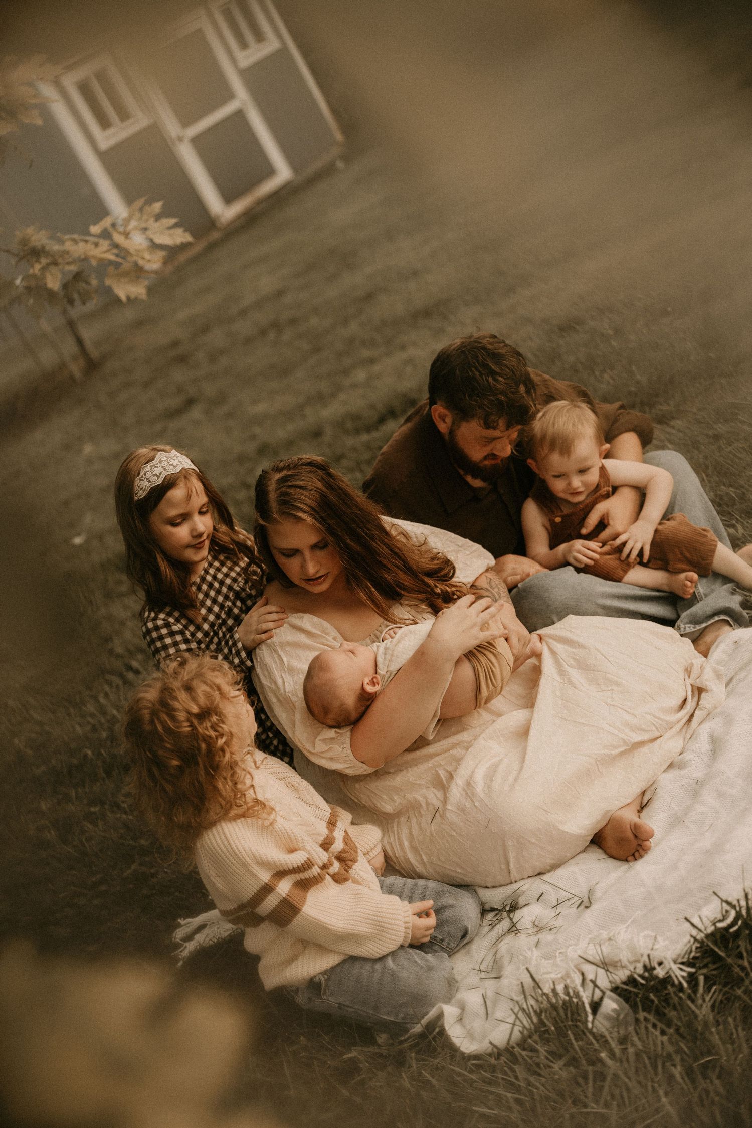 A family sits together on a blanket outdoors, mother cradling a newborn surrounded by children and partner.