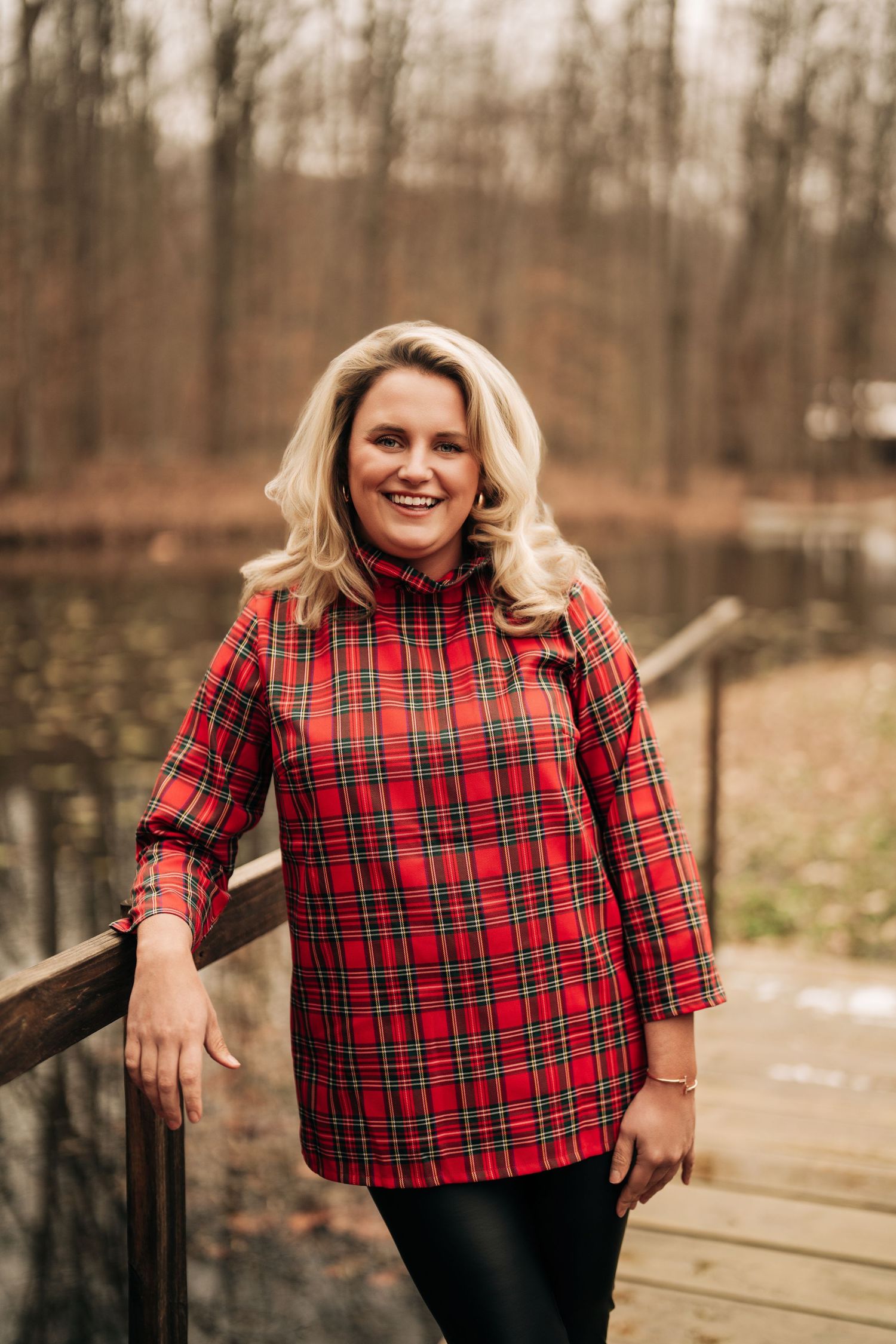 Woman in red plaid tunic smiles while standing on wooden deck in autumn forest setting.