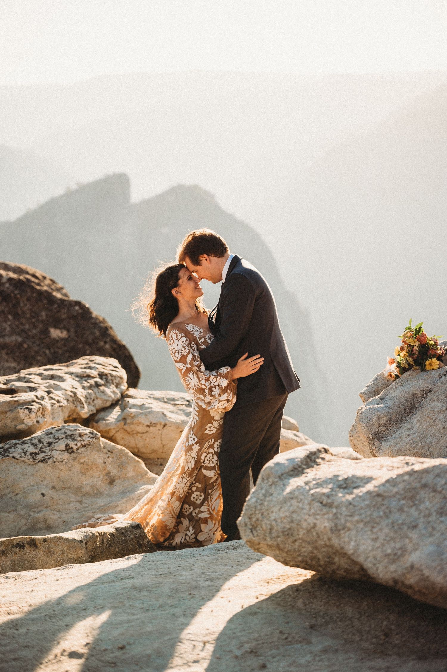 A couple embraces on a scenic mountain cliff during a romantic photoshoot at sunset.