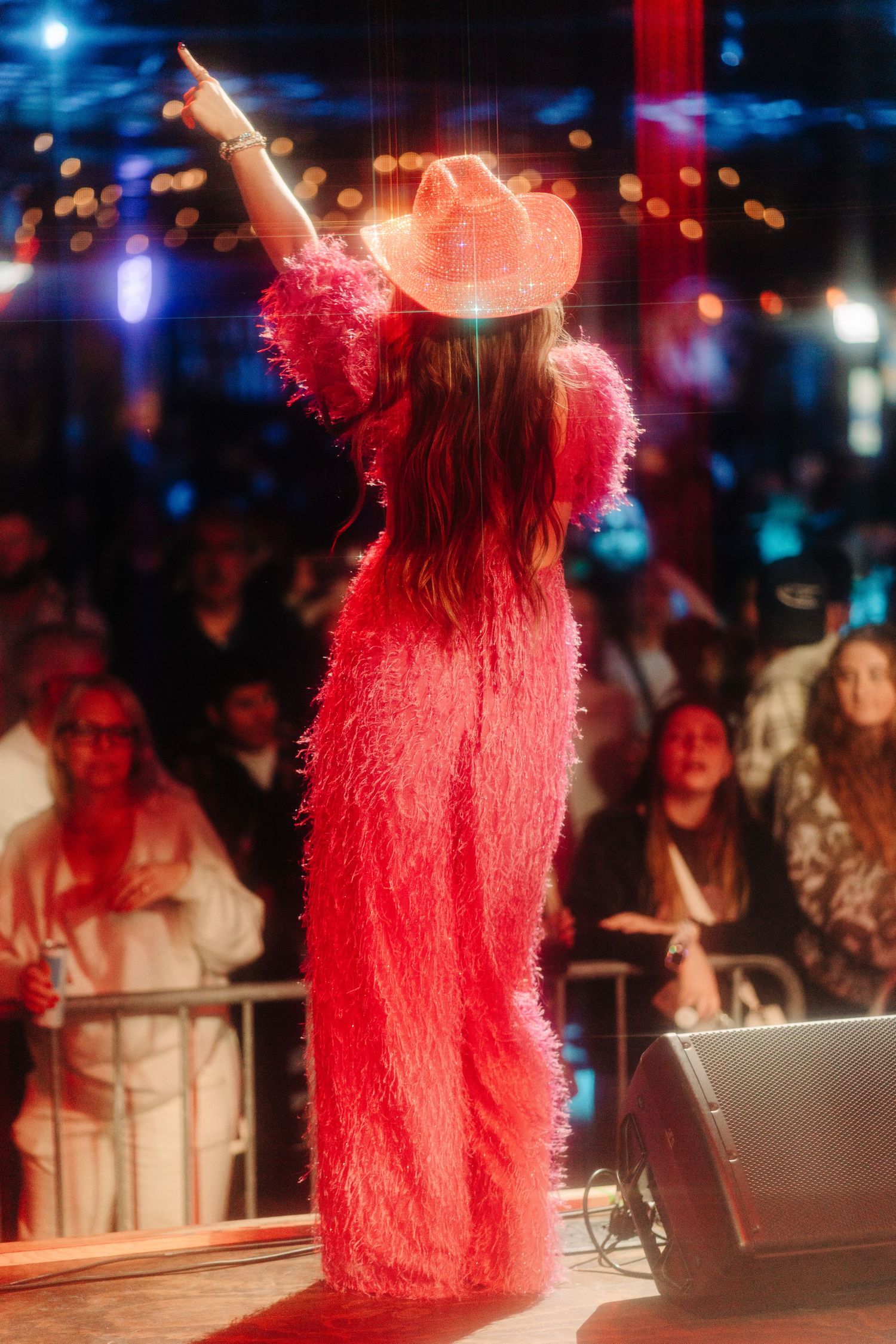 Performer in a bright pink furry costume dances energetically on stage with colorful lights and crowd in background.