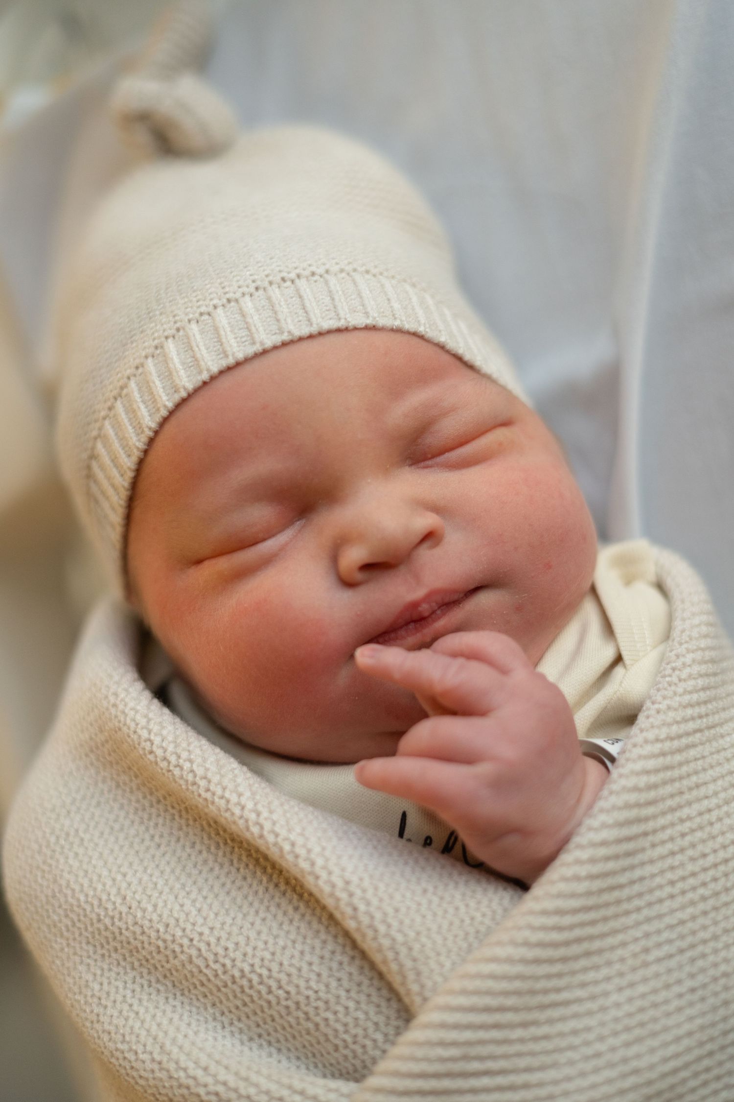 Sleeping newborn baby wearing cream knitted hat and blanket with peaceful expression.
