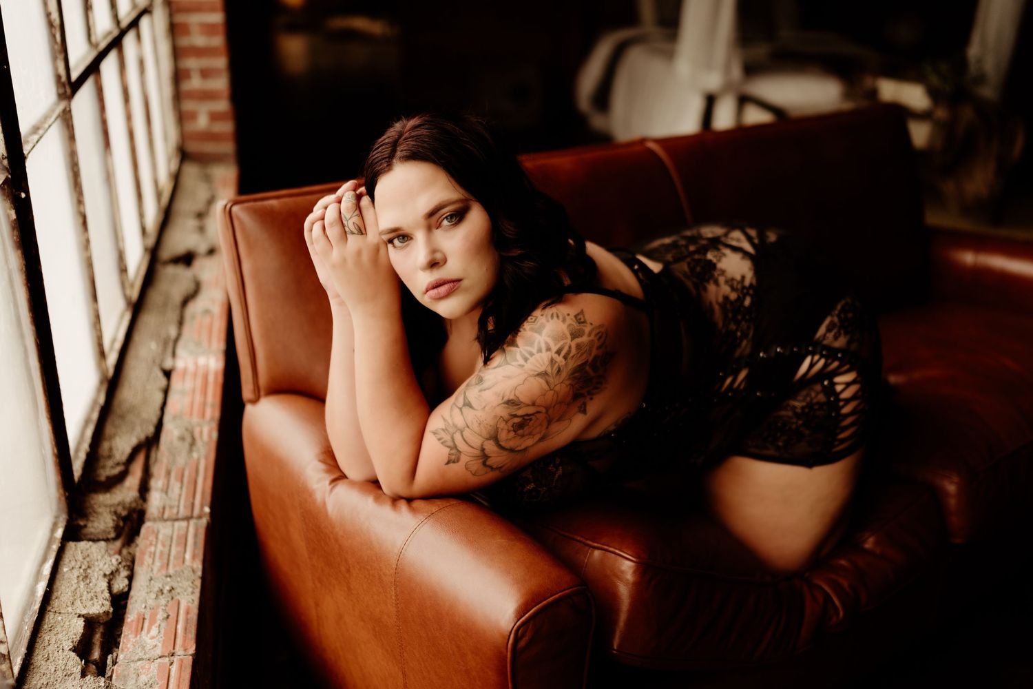Artistic portrait on leather couch near window with moody lighting.