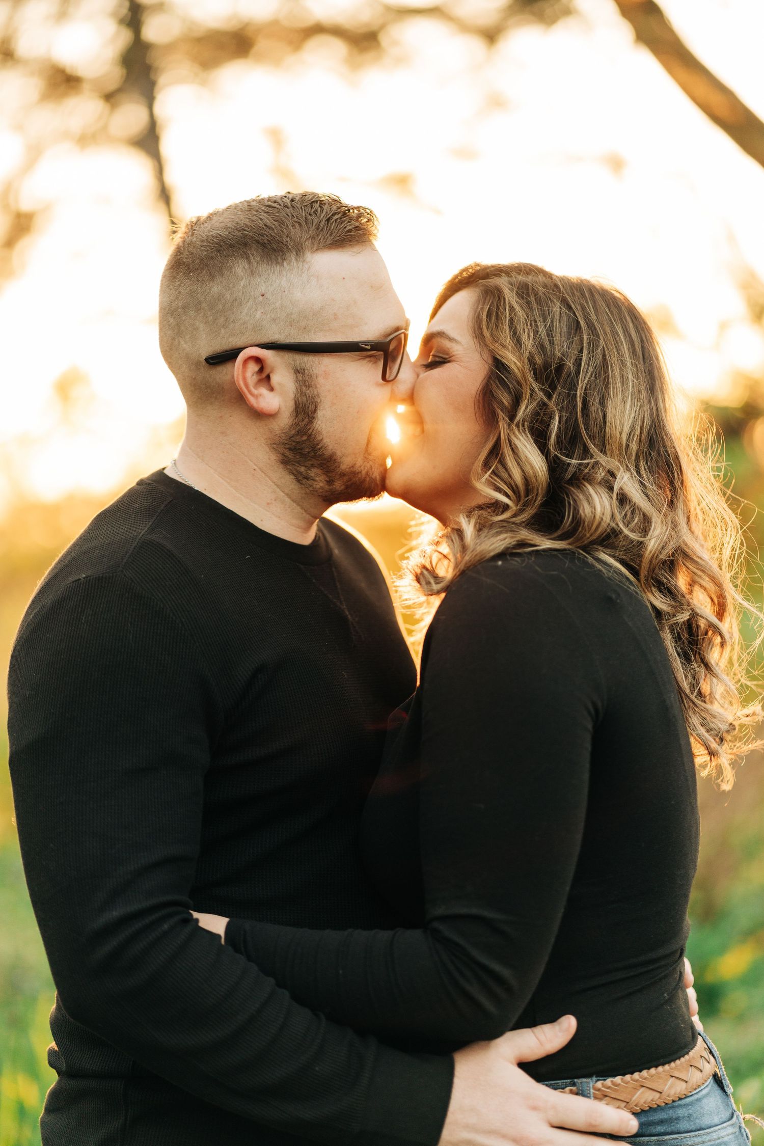 Romantic couple in matching black sweaters share a tender kiss at sunset in a sunlit outdoor setting.