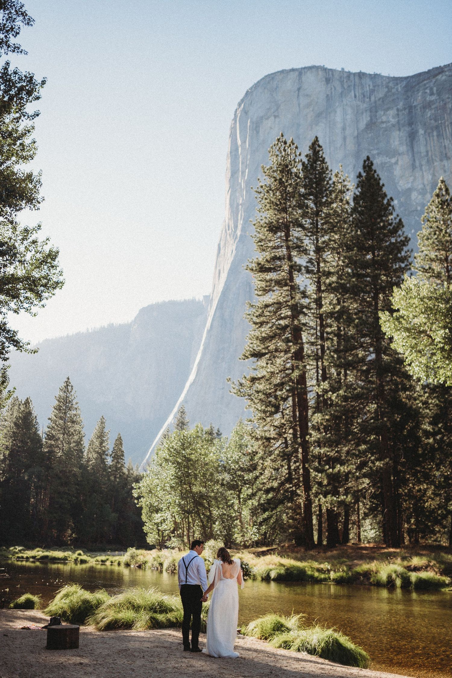 A wedding couple walks along a serene lake in Yosemite National Park with El Capitan towering in the background.