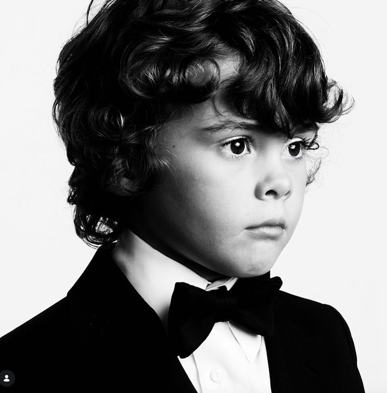 Black and white portrait of a young boy with curly dark hair wearing a black tuxedo jacket, white shirt and bow tie.