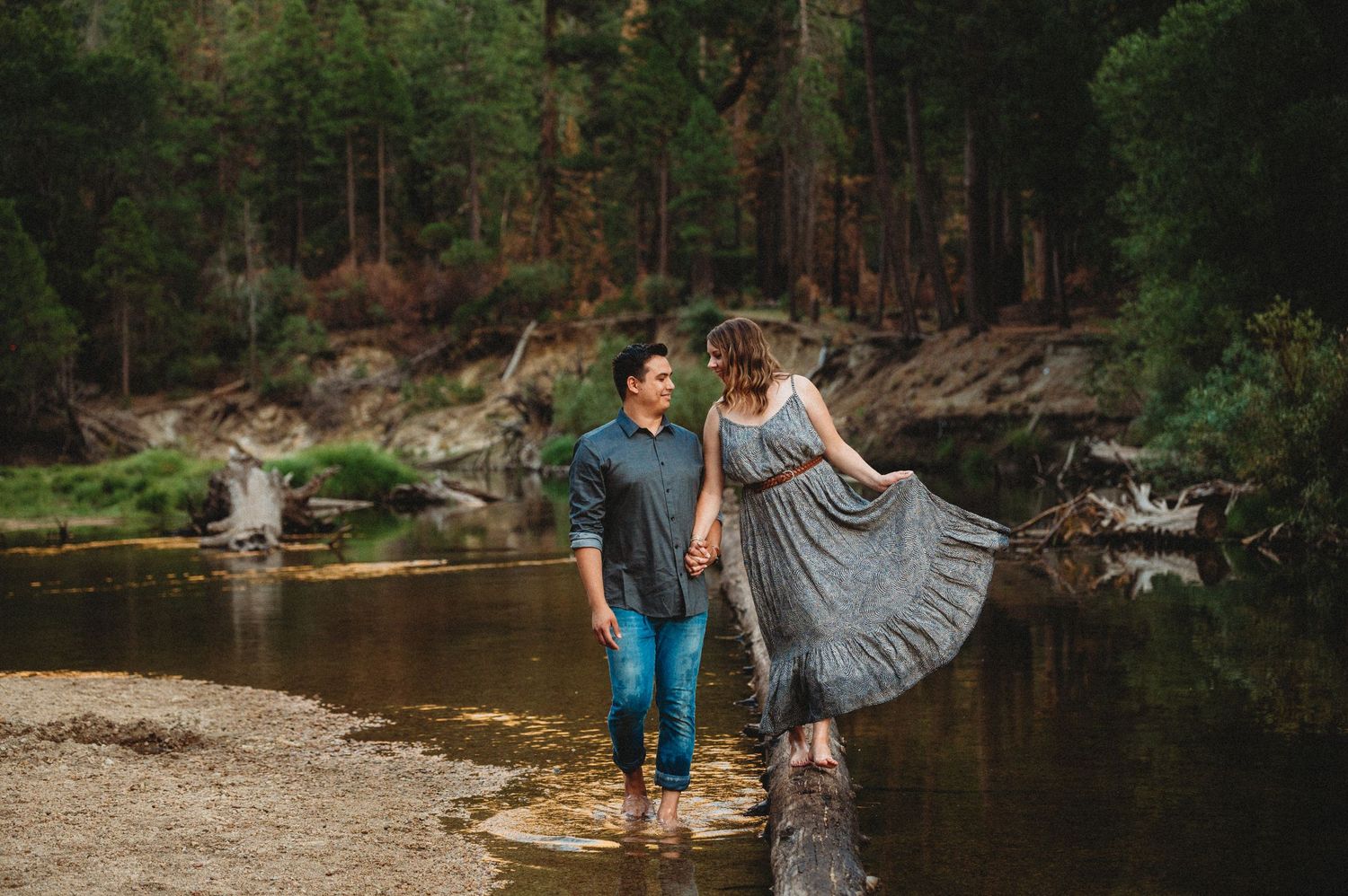 A couple stands on rocks by a serene forest lake, with the woman wearing a flowing gray dress.