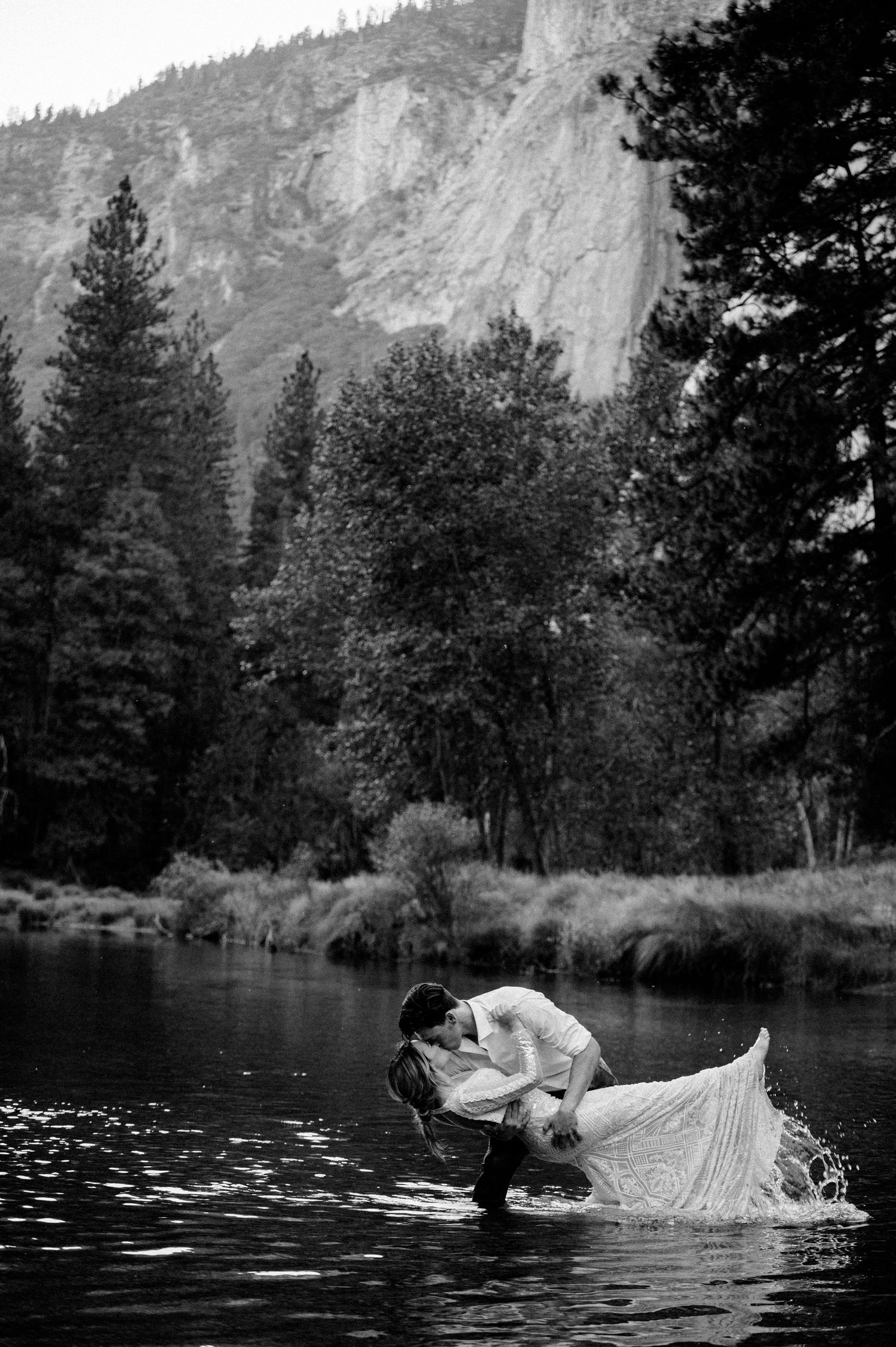 A romantic couple embraces in a lake with dramatic granite cliffs and evergreen forest of Yosemite Valley in background.