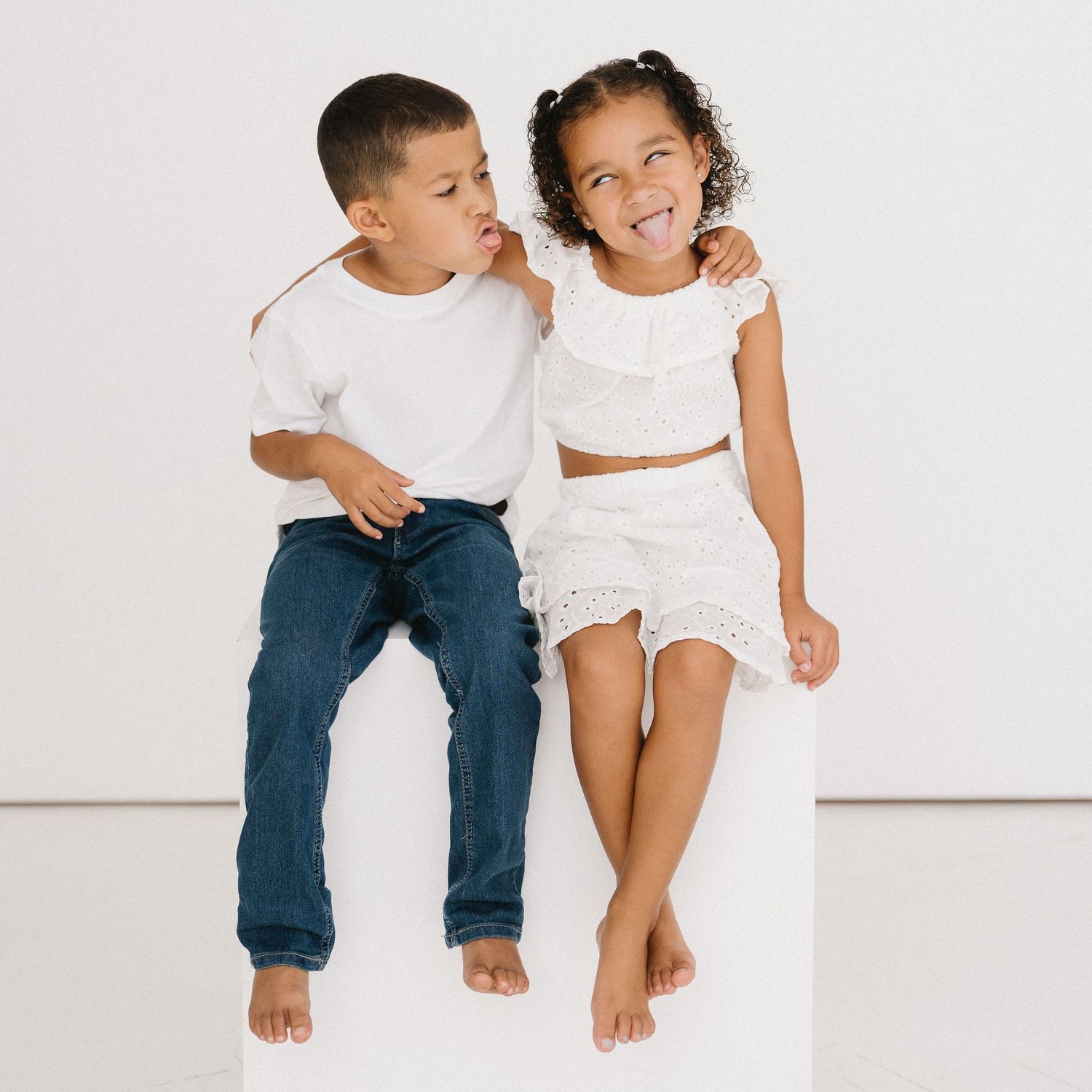 Two children in casual white outfits sitting together against a white background, sharing a playful moment.
