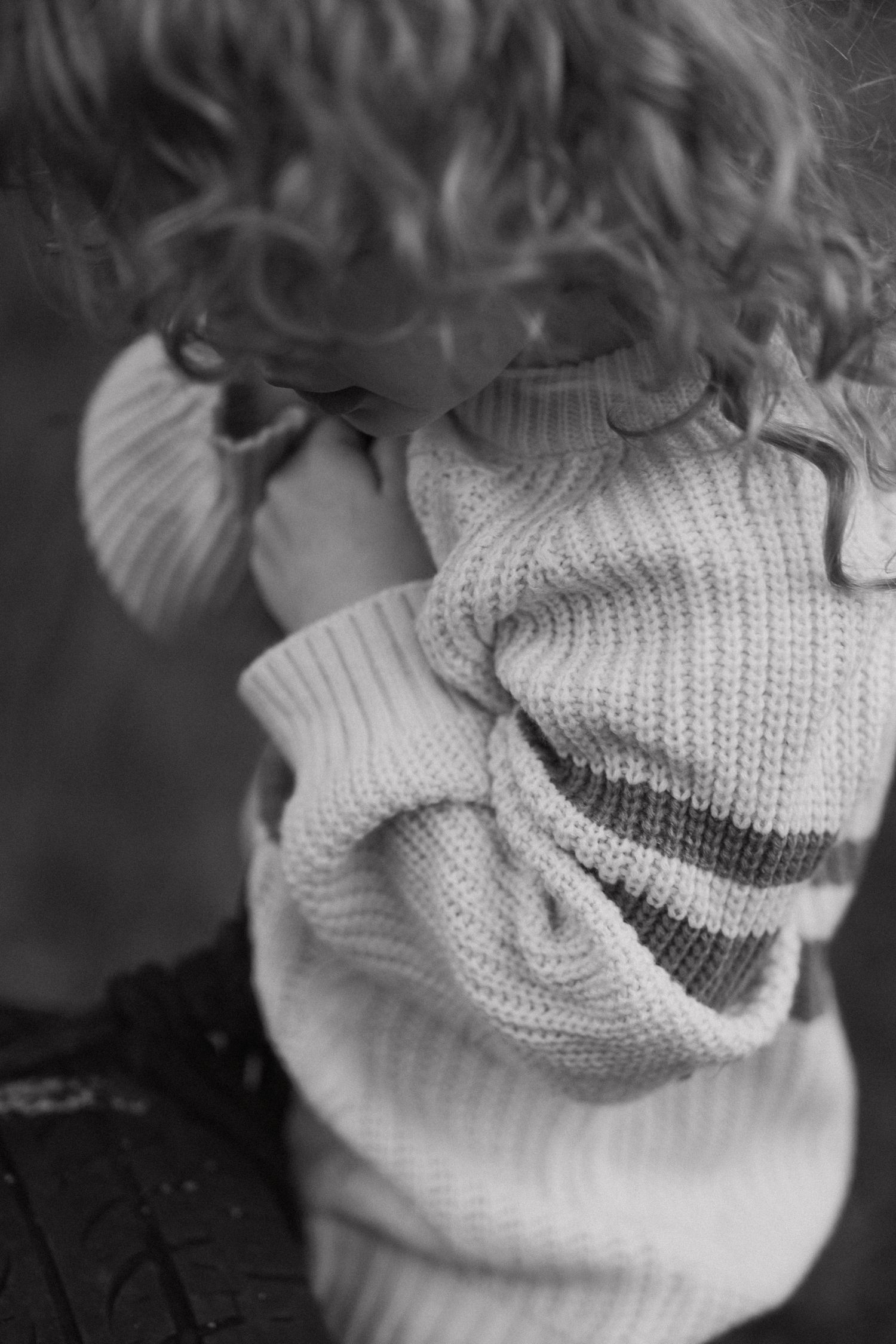 Black and white photo of a person with curly hair wearing an oversized knit striped sweater, head tilted down.