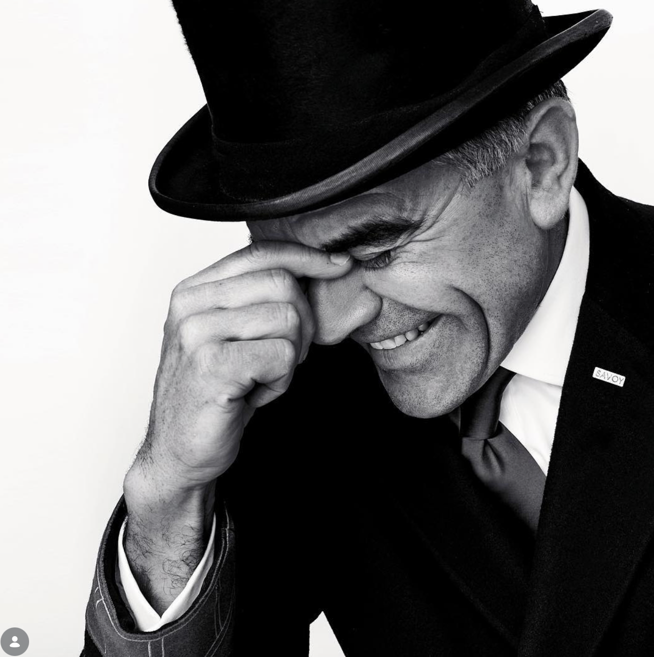Black and white portrait of a smiling man in a top hat and dark suit, laughing with his hand raised to his eye.