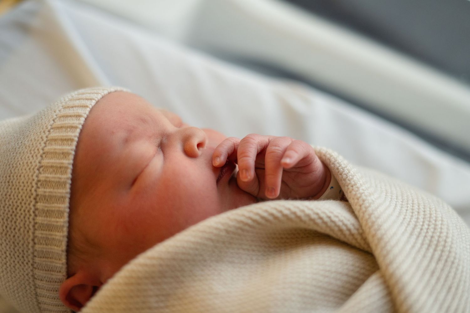 Newborn baby yawning while wrapped in cream-colored blanket on white bedding.