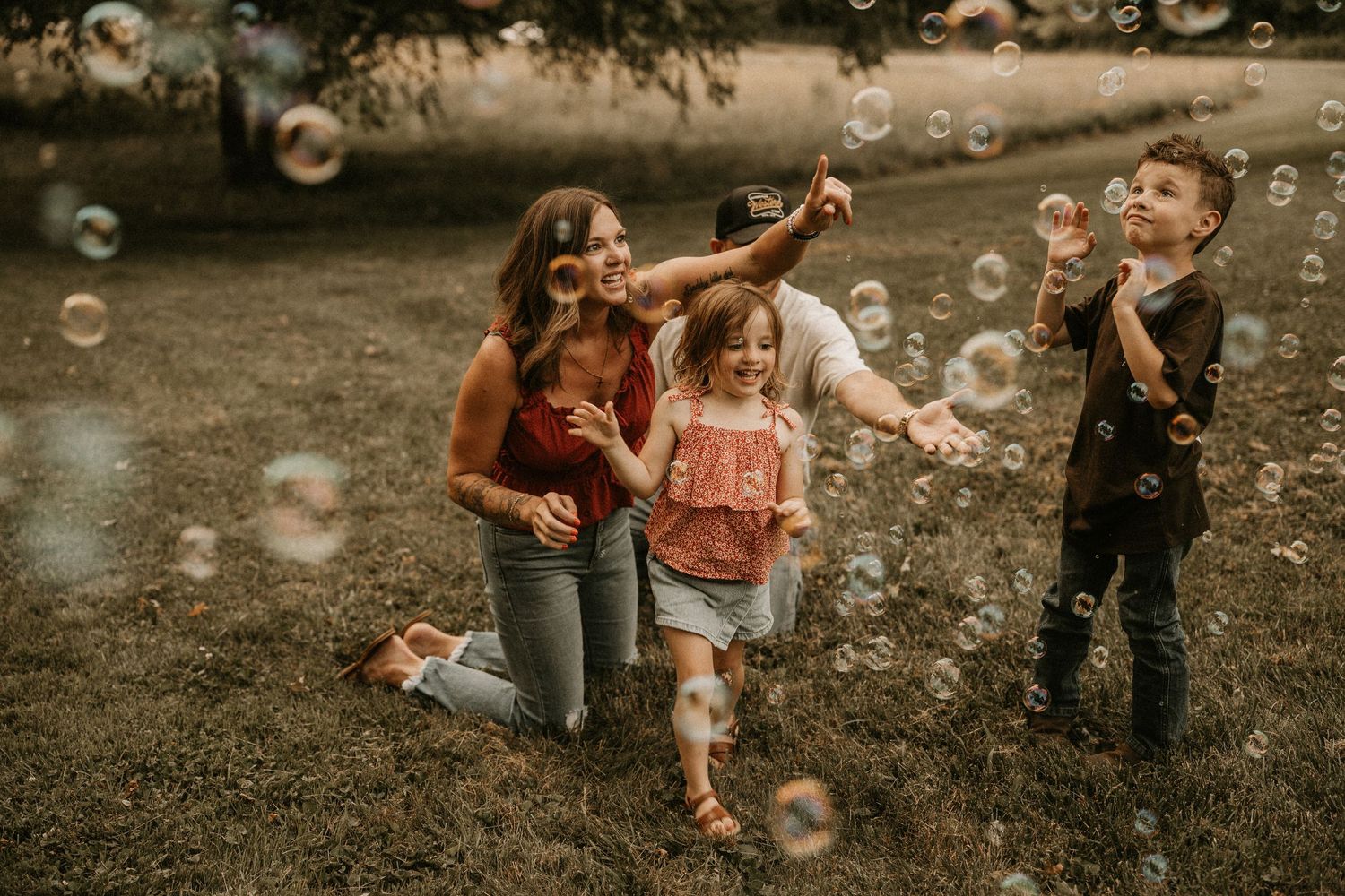 Family playing with bubbles outdoors on grass, mom kneeling with two young children laughing and reaching for floating soap bubbles.