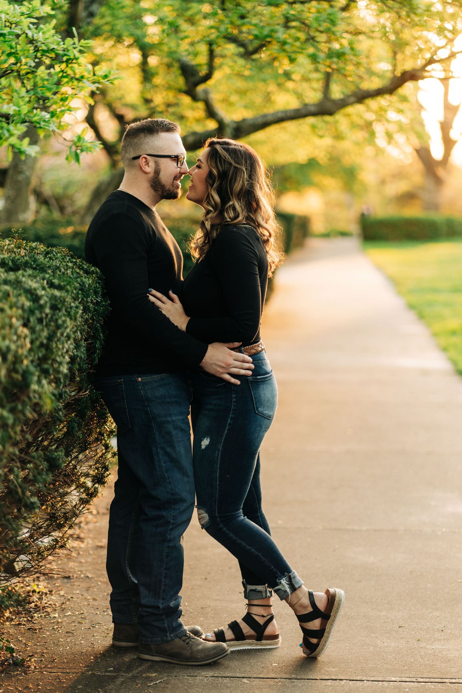 A couple embraces on a sunlit path surrounded by lush greenery in a park-like setting.