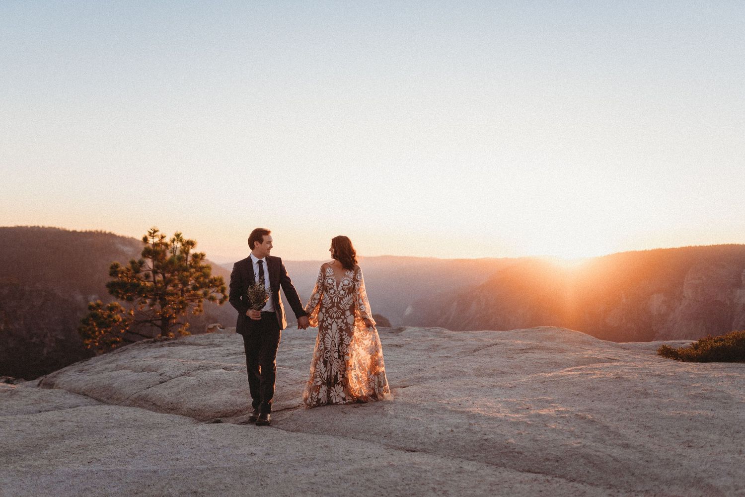 A couple in wedding attire walks hand in hand on a mountain cliff during golden hour sunset.