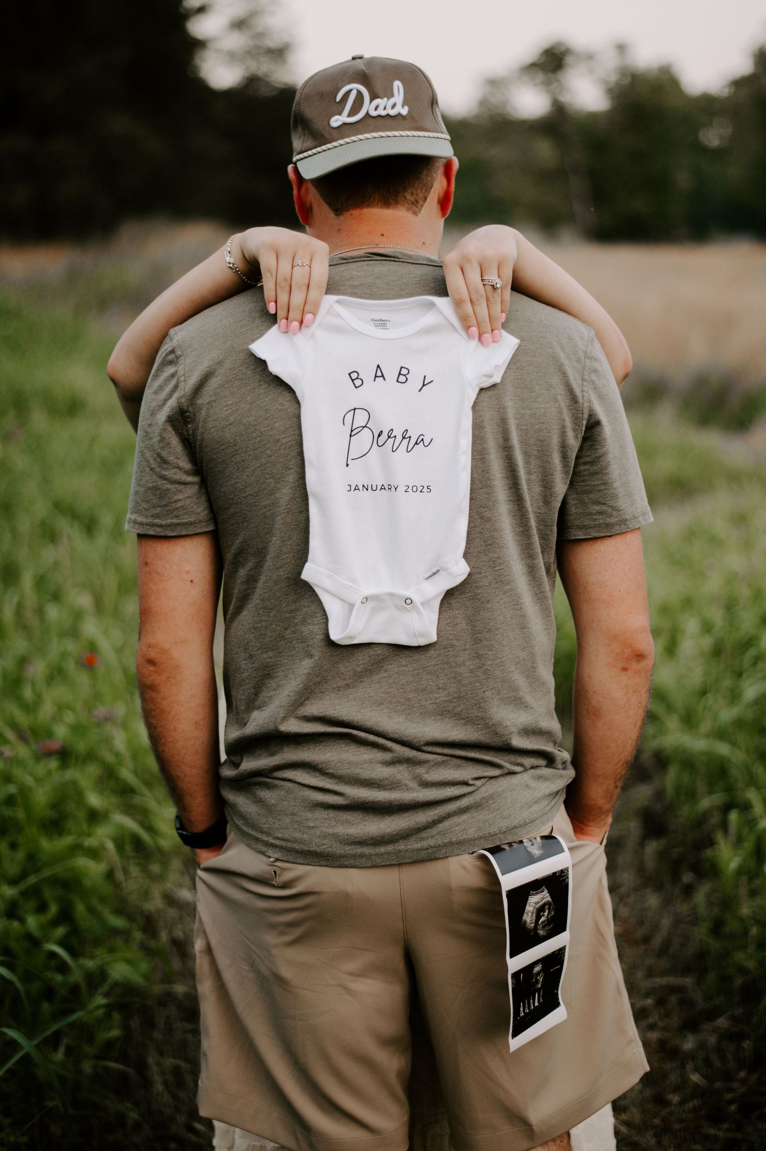 Person in grey shirt and cap holds baby onesie while standing in grassy field with ultrasound photos.
