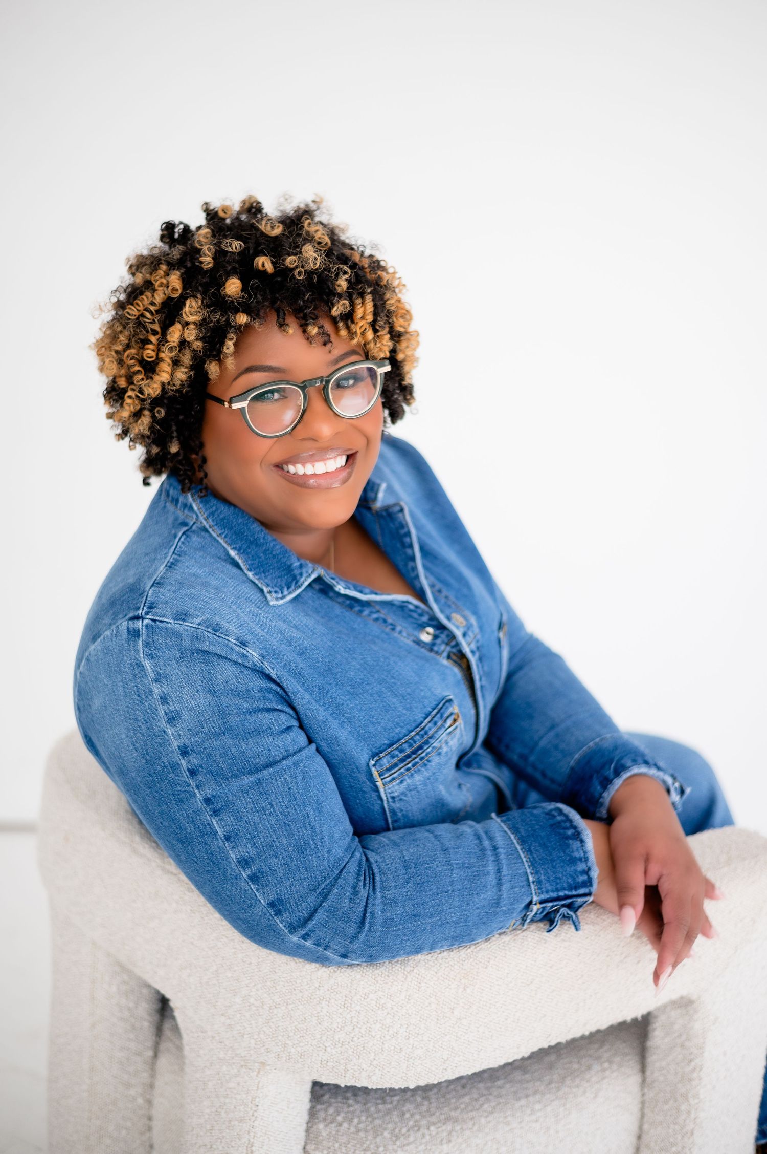 Person with curly blonde hair and glasses relaxes on white chair wearing denim jacket and smiling warmly.