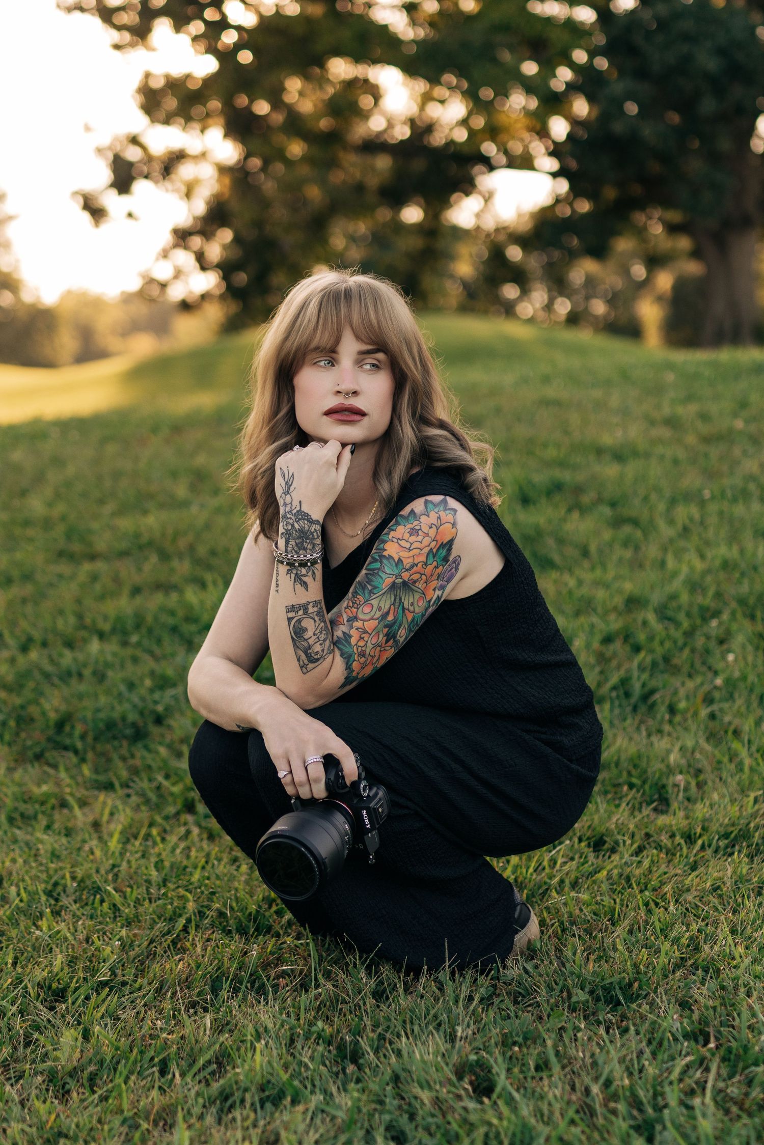 Tattooed woman in black outfit sitting on grass holding a camera, posing outdoors at golden hour.