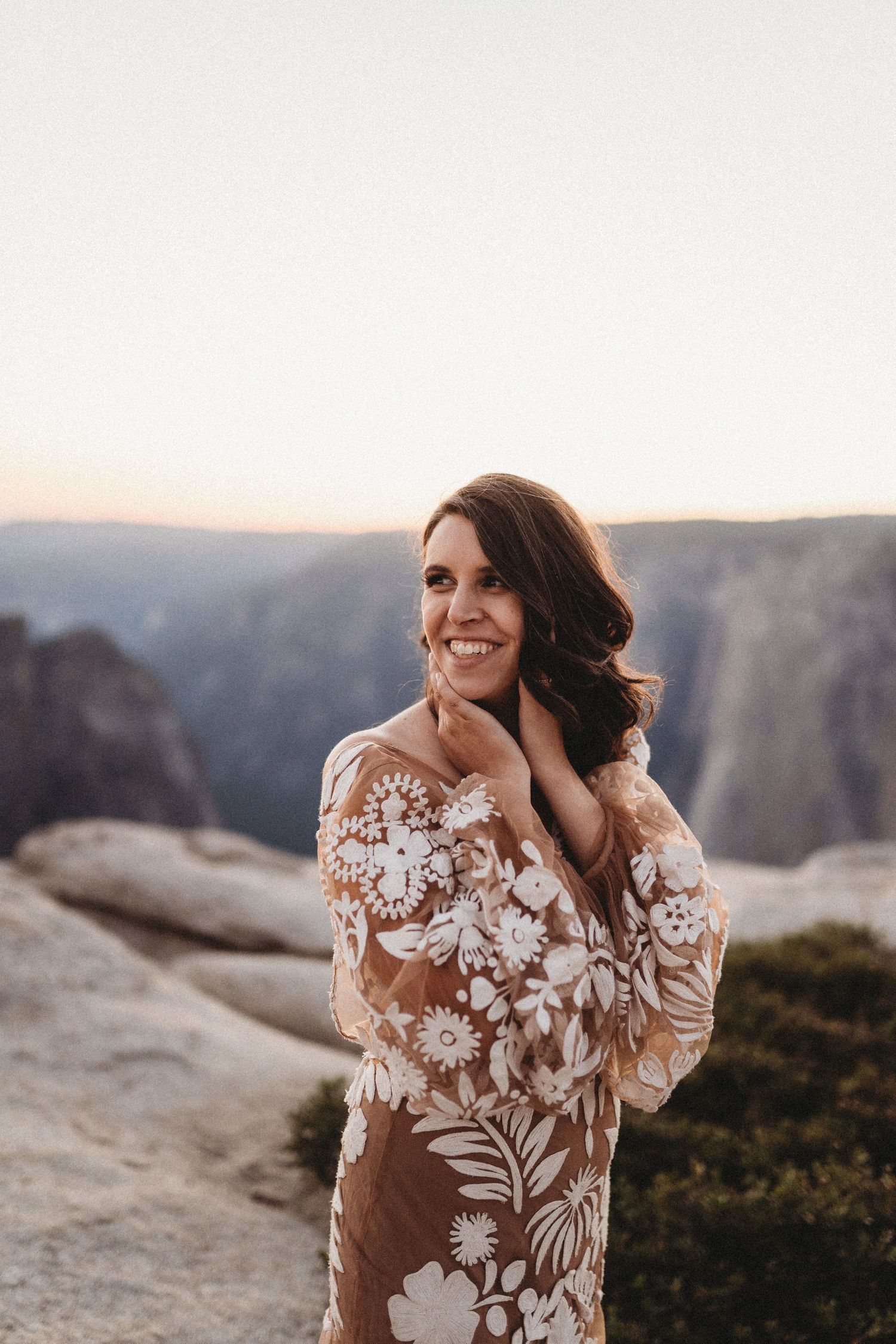 A floral lace wedding dress with sheer details photographed against a dramatic mountain landscape at sunset.