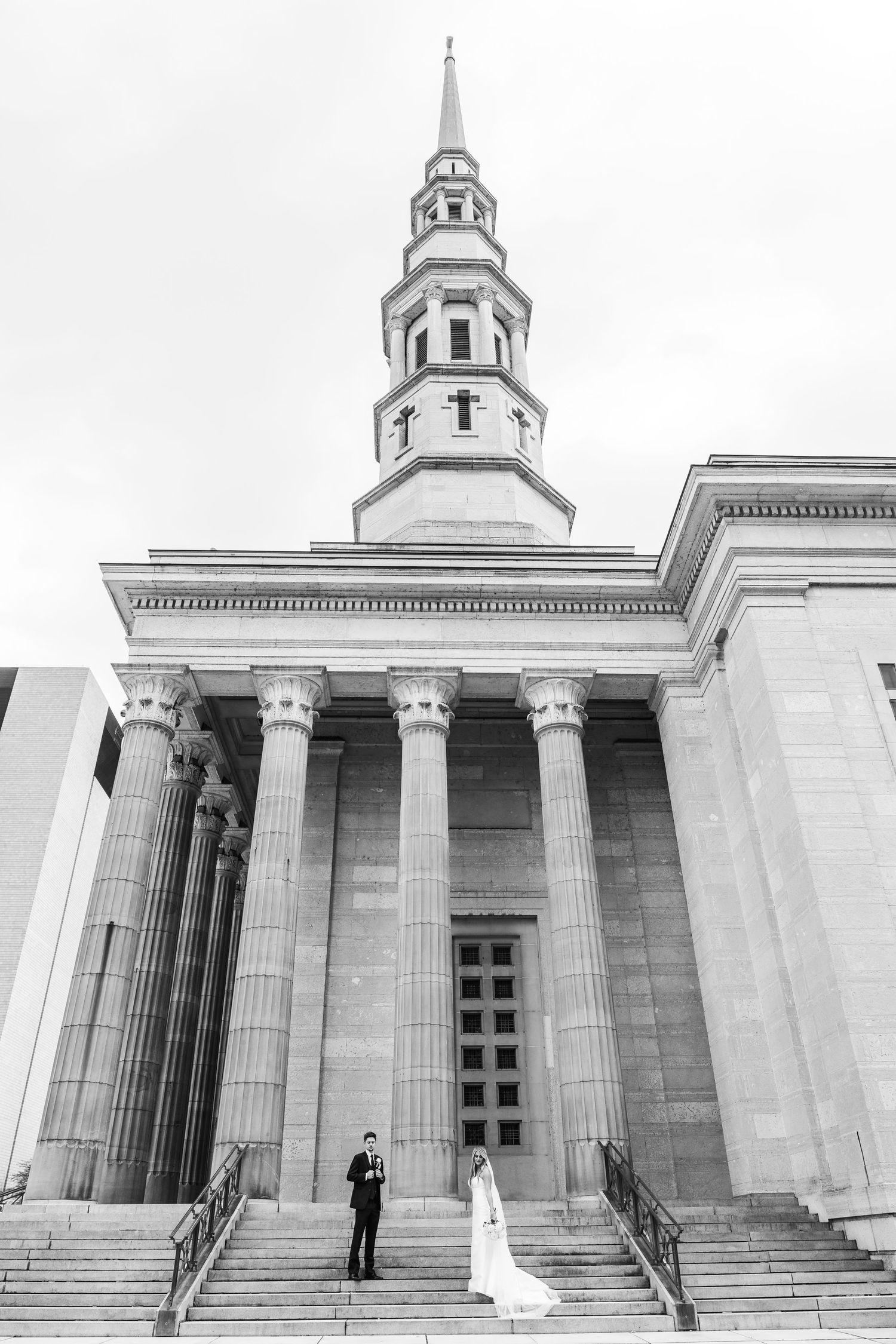 A black and white photo of a grand church with classical columns and a tall steeple where a couple stands on the steps.
