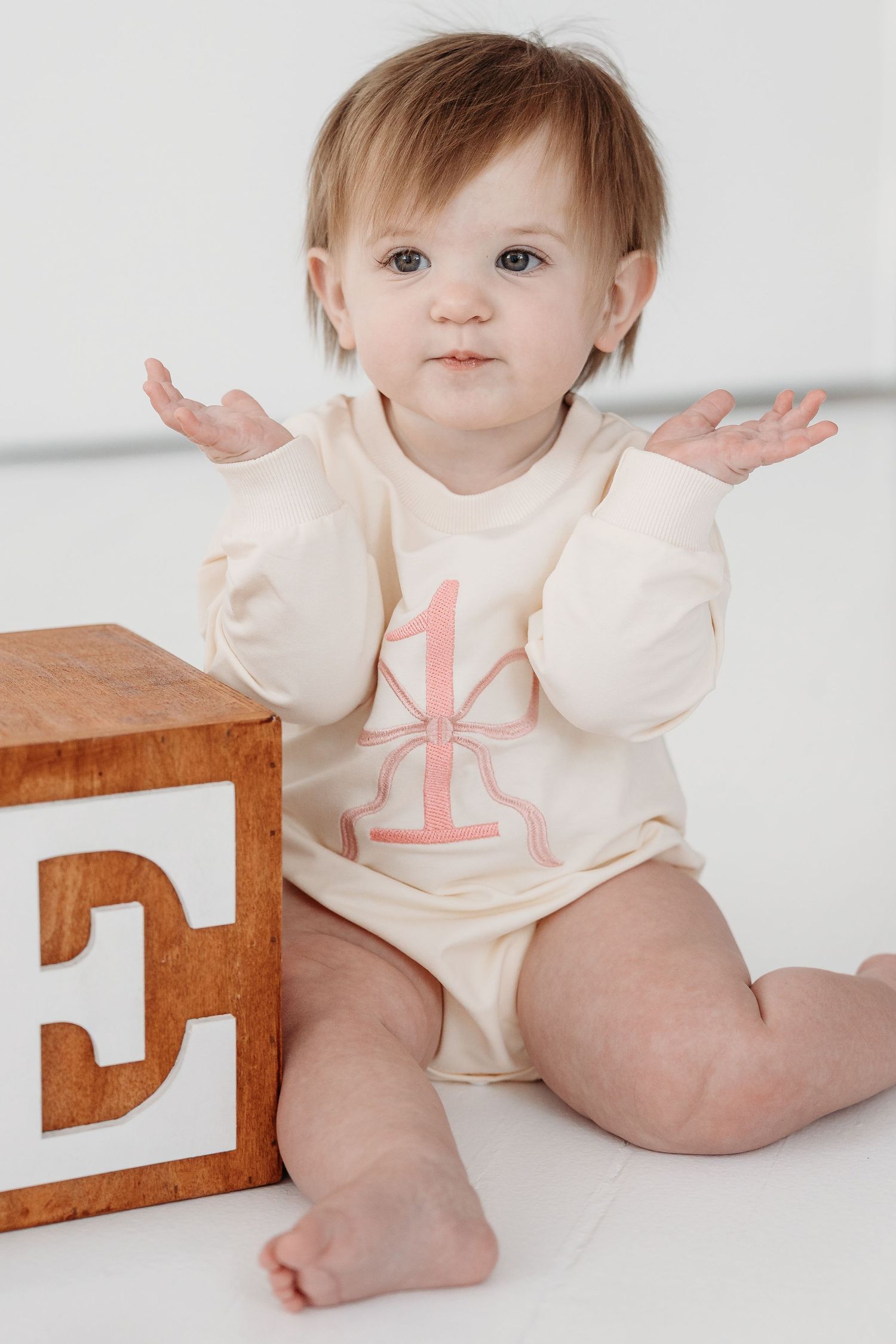 Baby in cream colored onesie with number 1 design sits next to wooden letter E block on white background.