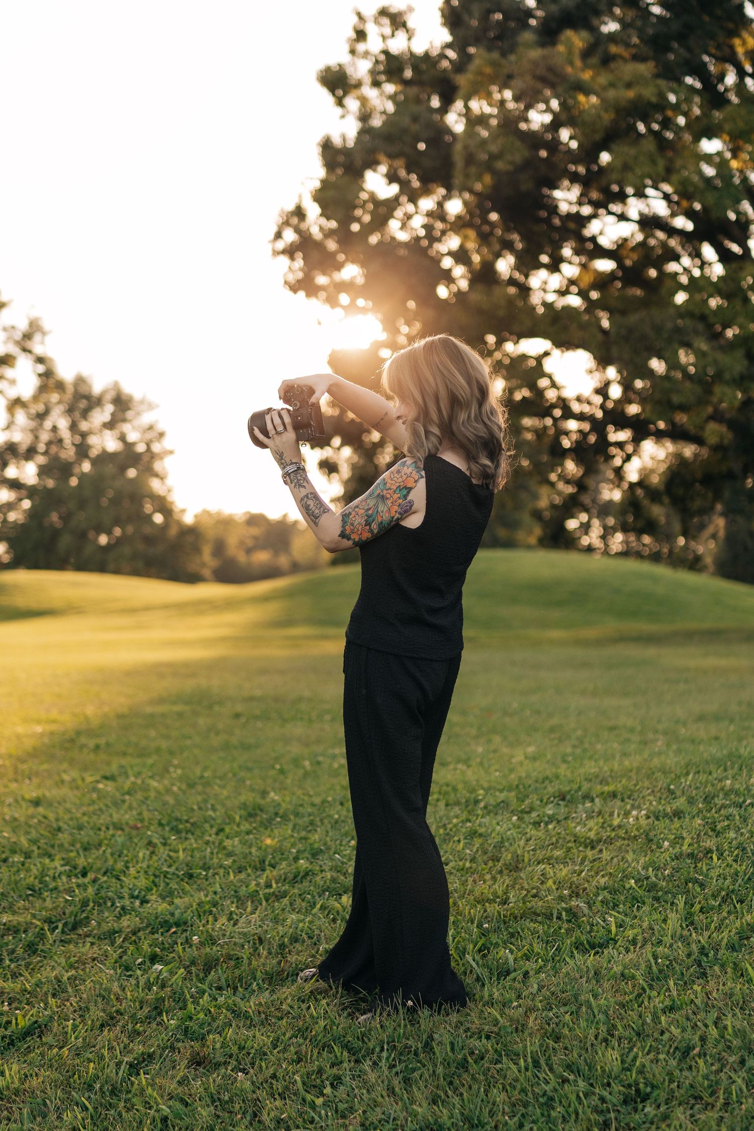 Tattooed woman in black dress photographing outdoors at golden hour sunset in lush green park.