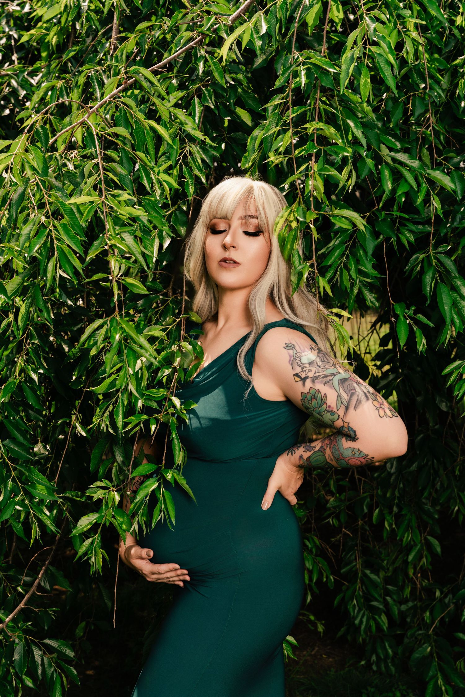 Person in emerald green dress poses against lush green foliage background with visible arm tattoo.