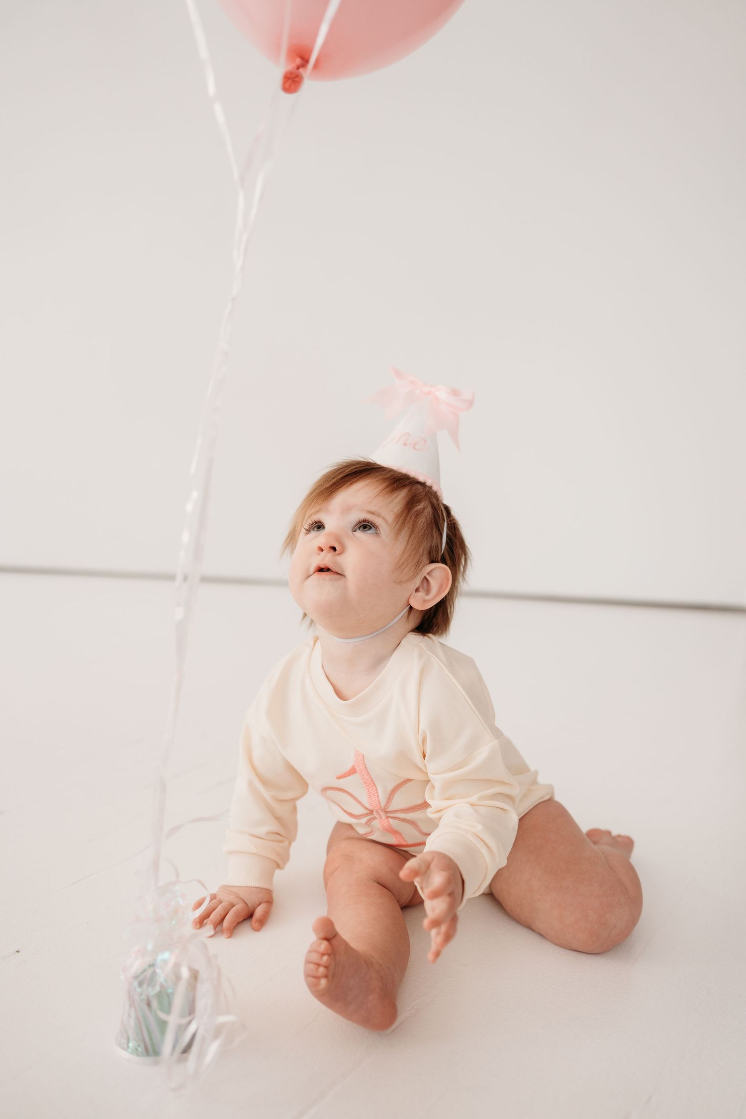A baby in a cream-colored outfit lies on a white floor looking up at pink balloons floating above.