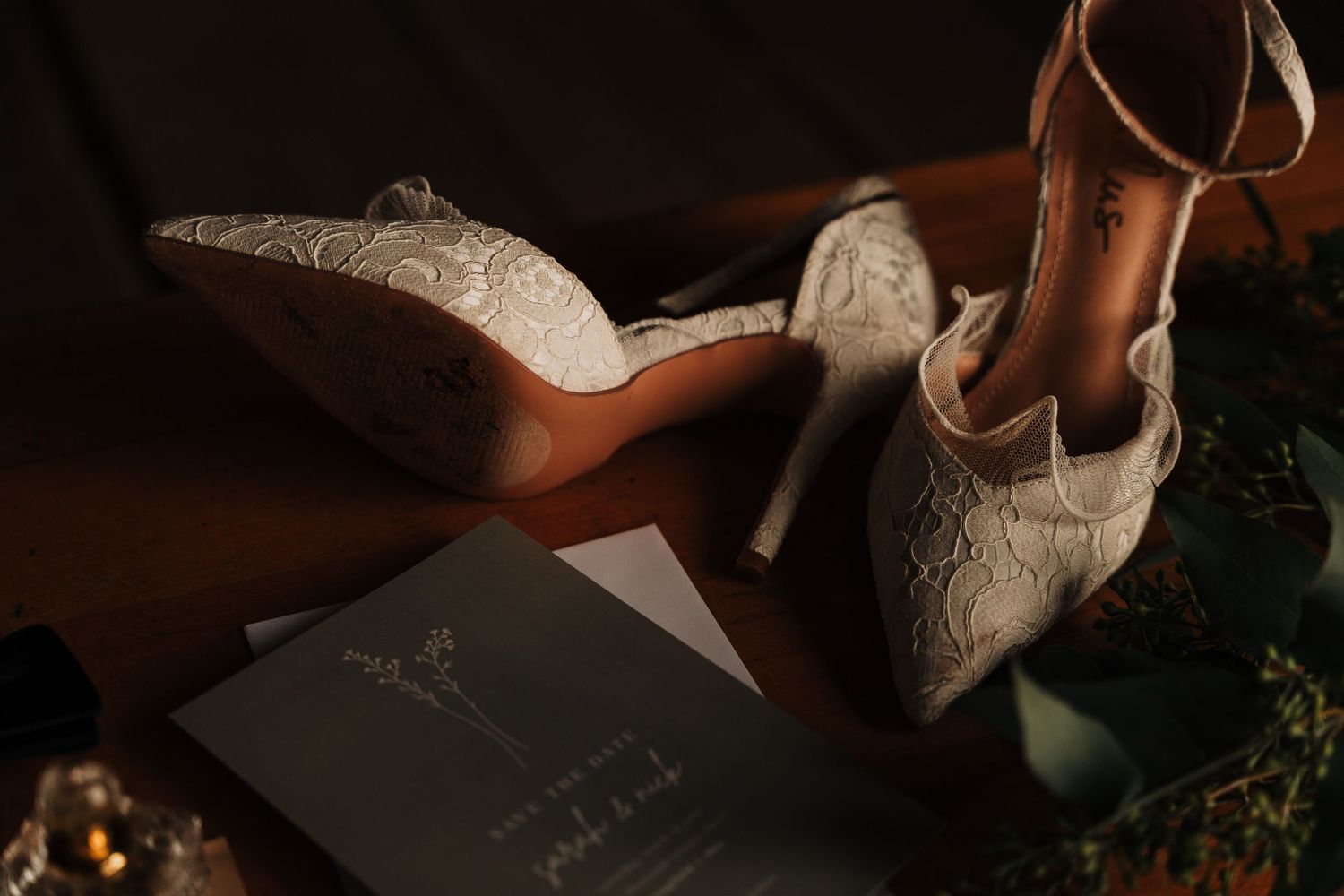 Elegant white lace bridal heels and a dark wedding invitation card on a moody background.