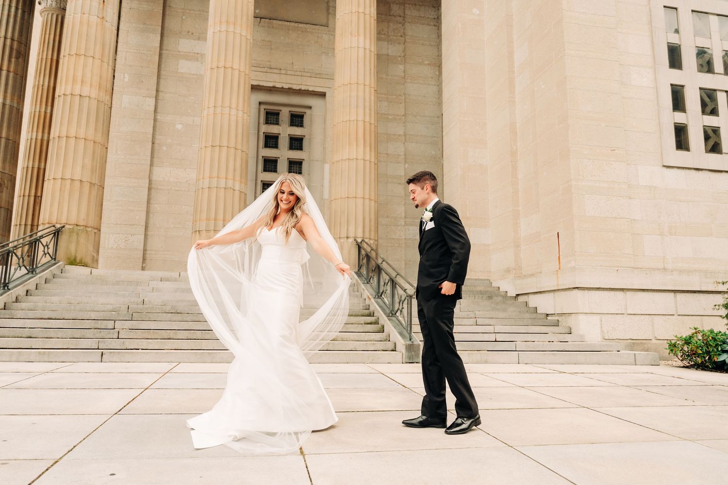 A wedding couple poses for romantic photos on the grand steps of a neoclassical stone building.