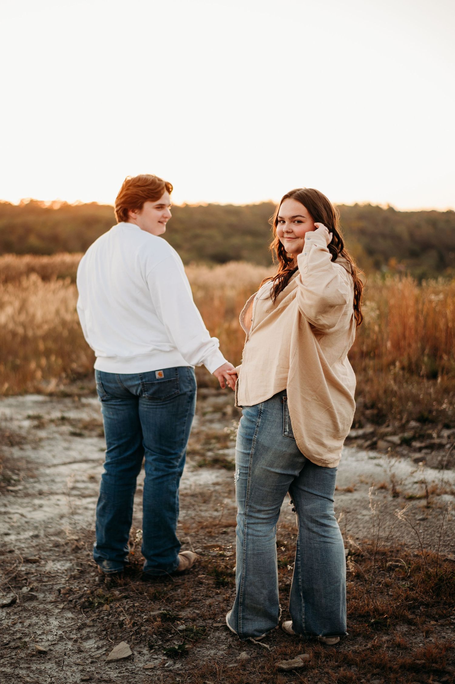 Couple holding hands in a field at sunset, woman in beige sweater touching her hair.