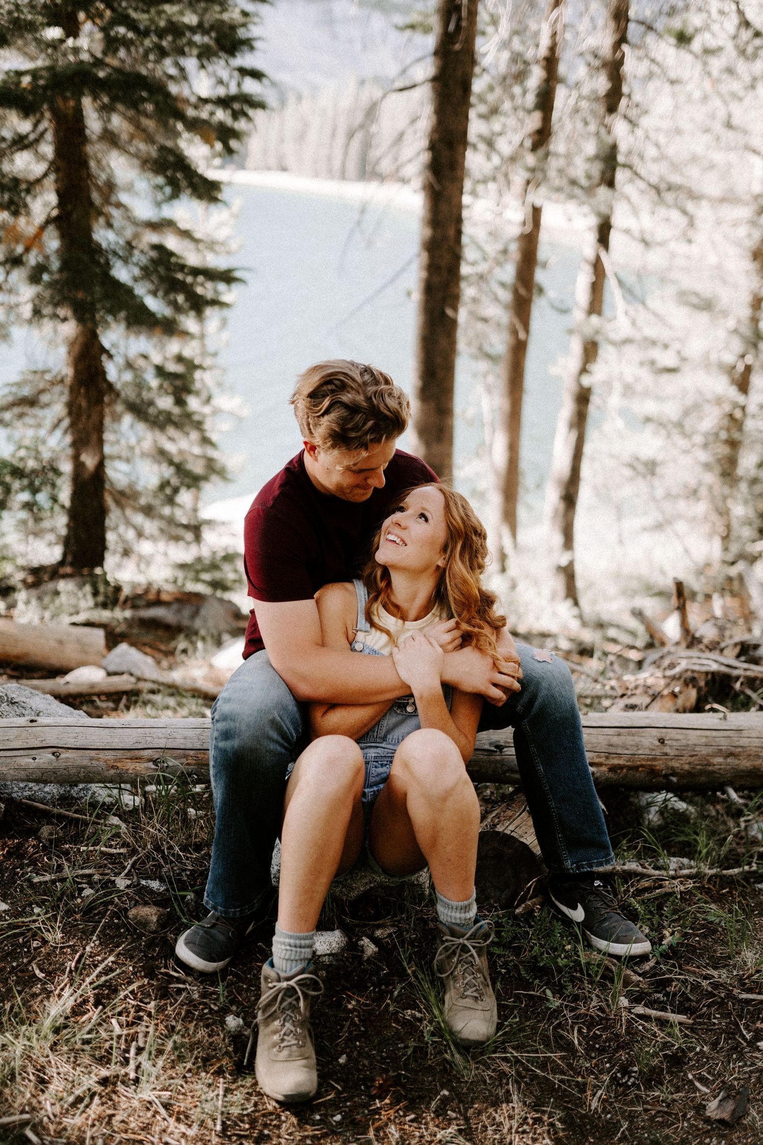 A couple embraces on a log in a forest setting with a waterfall visible in the background.