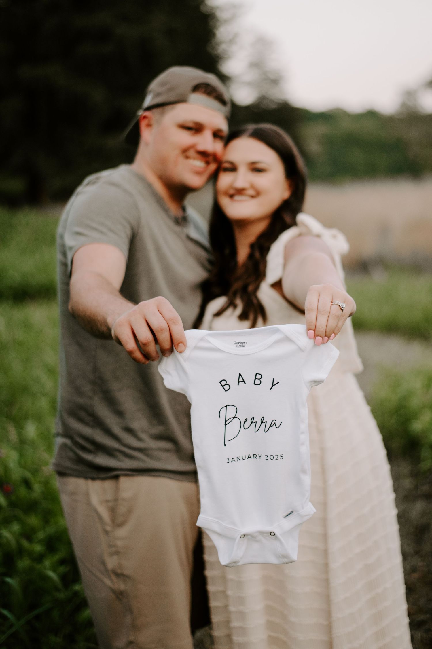 A couple holds up a white baby onesie with text announcing a January 2024 pregnancy in an outdoor setting.