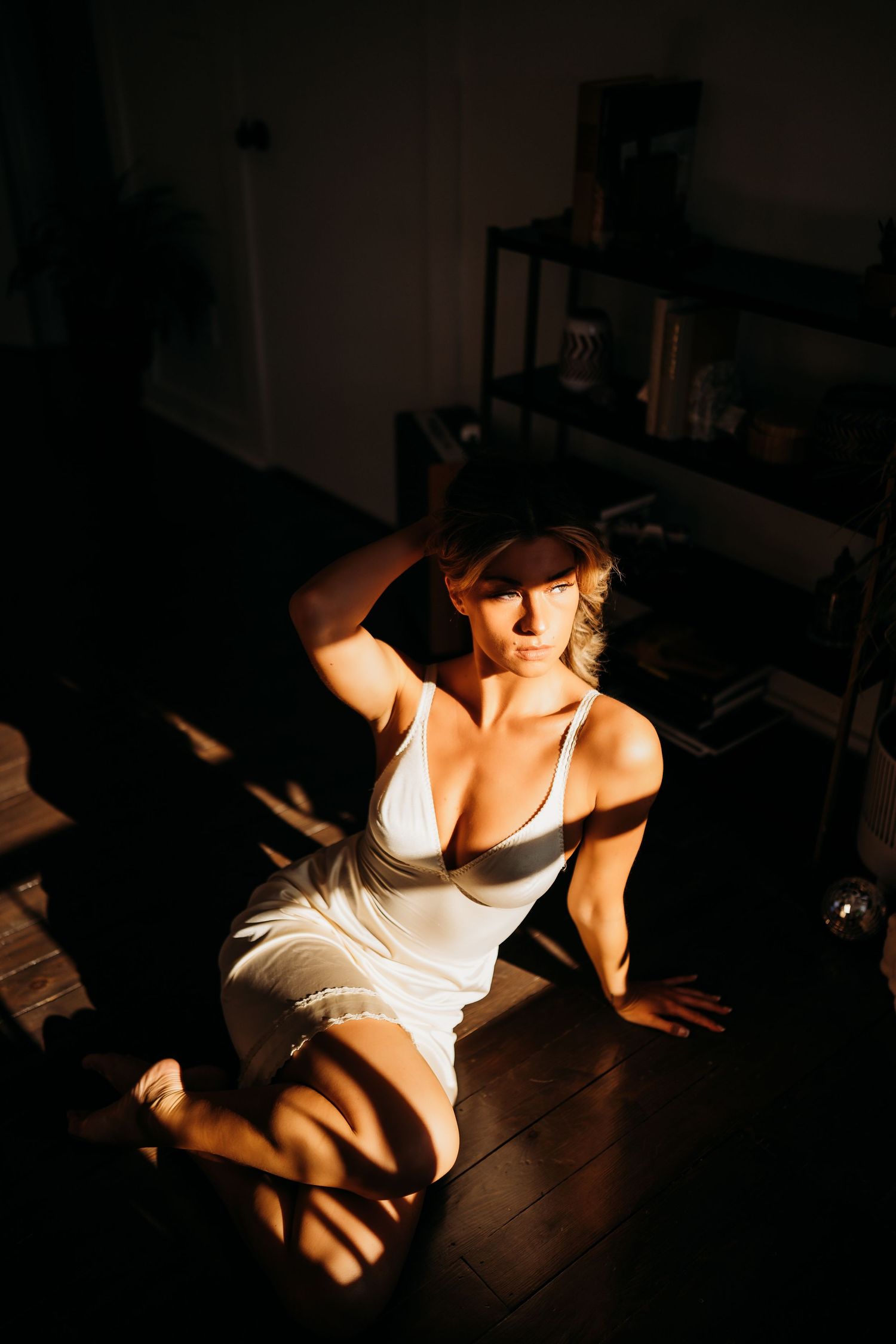 Artistic boudoir photograph in a dim room with dramatic shadows and warm lighting on hardwood floors.