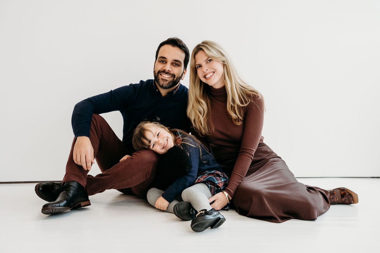 Happy family of three sitting on white studio floor, parents smiling with young child leaning on dad.