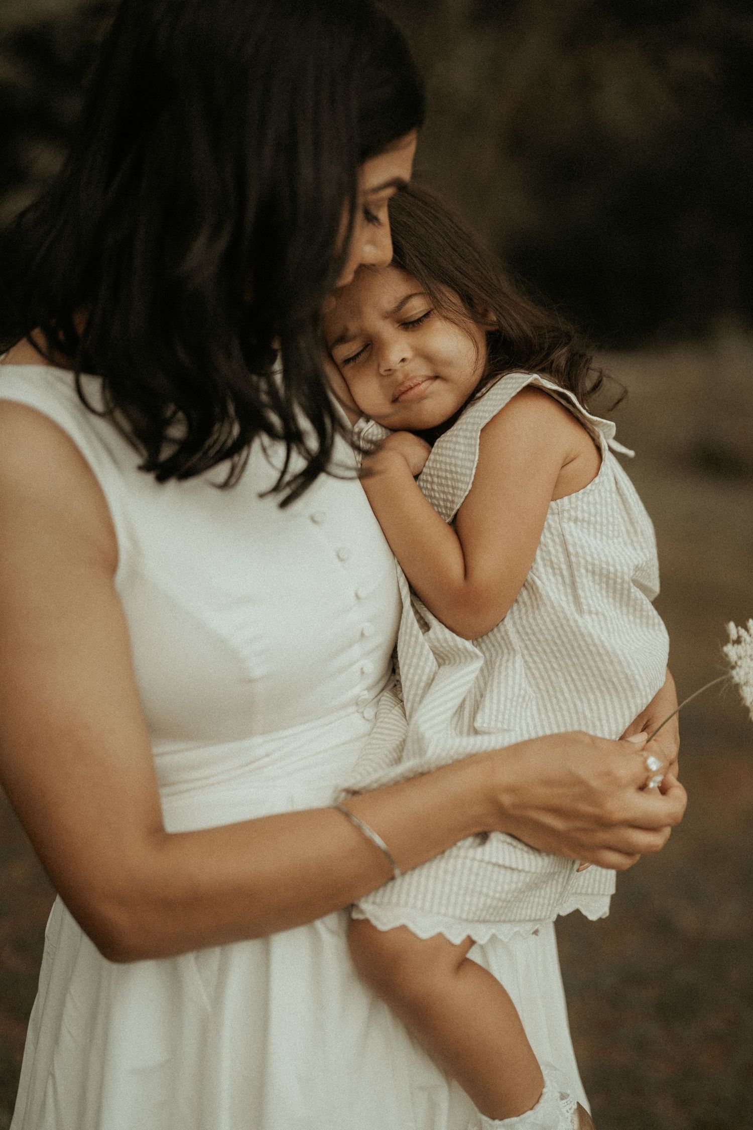 A mother in a white dress tenderly holds her young daughter, who rests her head on her chest with eyes closed.
