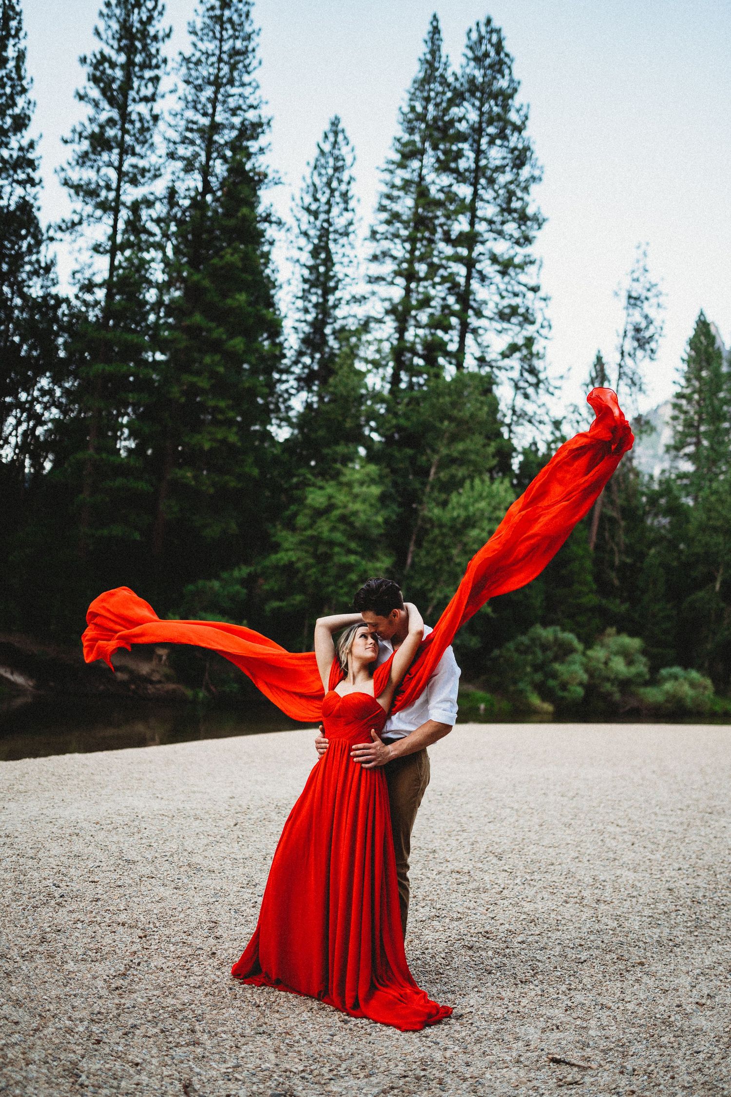 Woman in flowing red dress twirls gracefully in a forest clearing surrounded by tall pine trees.