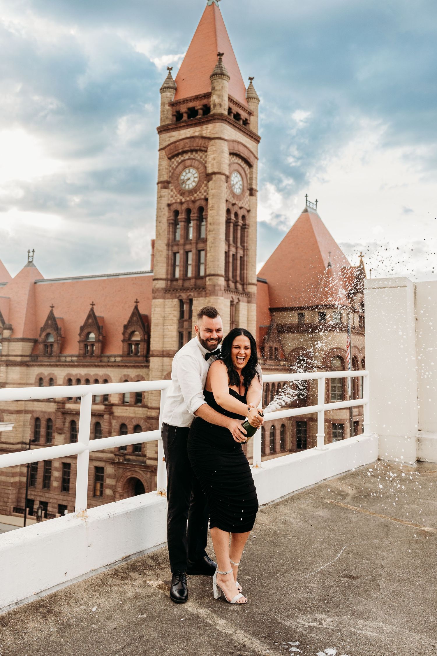 A couple poses together on a rooftop terrace with historic brick architecture and clock towers in the background.