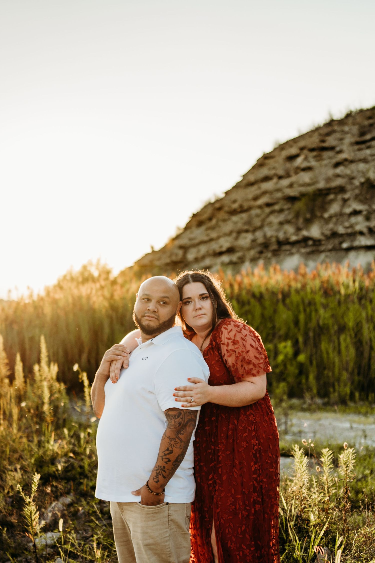 Couple embraces in golden sunlight against rocky cliffside during outdoor photo session at sunset.