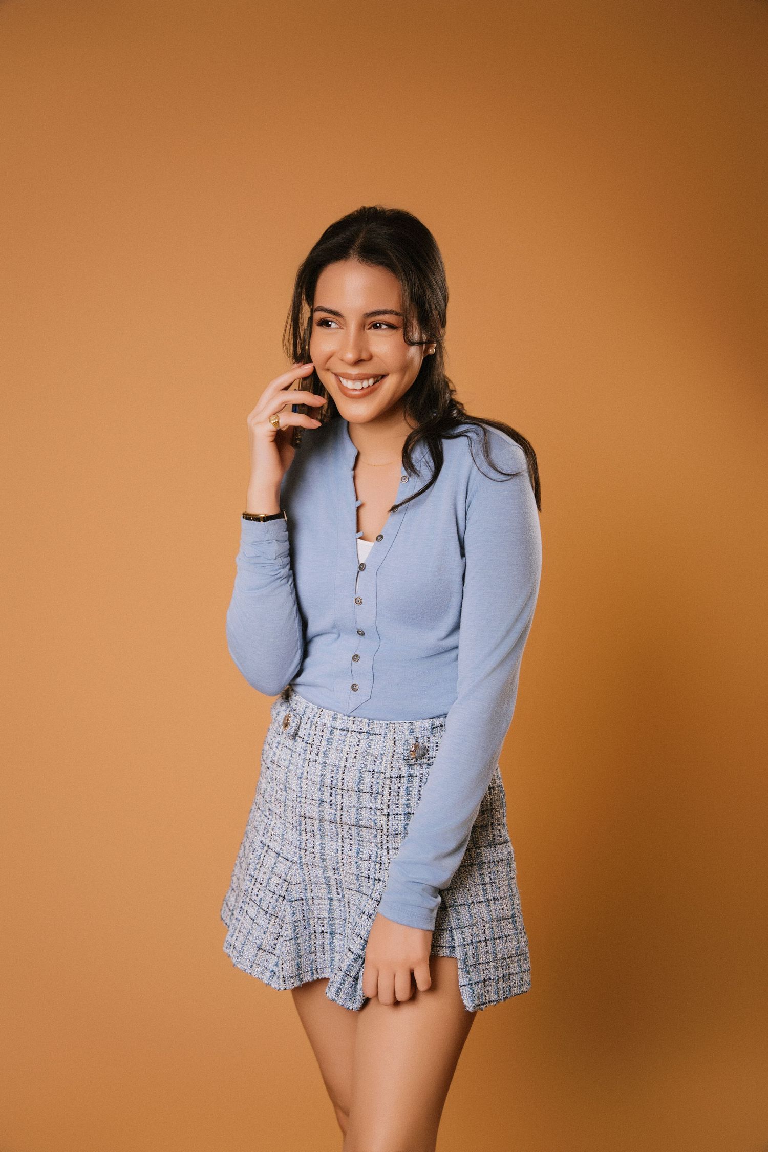 Young woman in light blue cardigan and plaid skirt smiling while talking on phone against orange background.