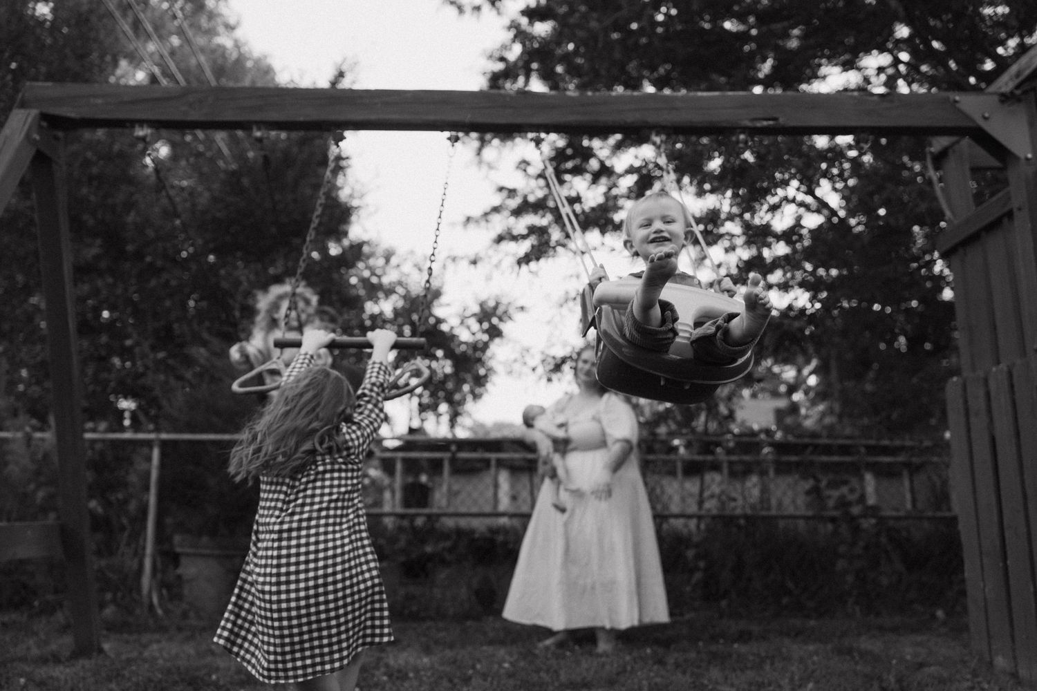 Black and white photo of children playing on a backyard swing set, with a girl in a dress watching a child swing.