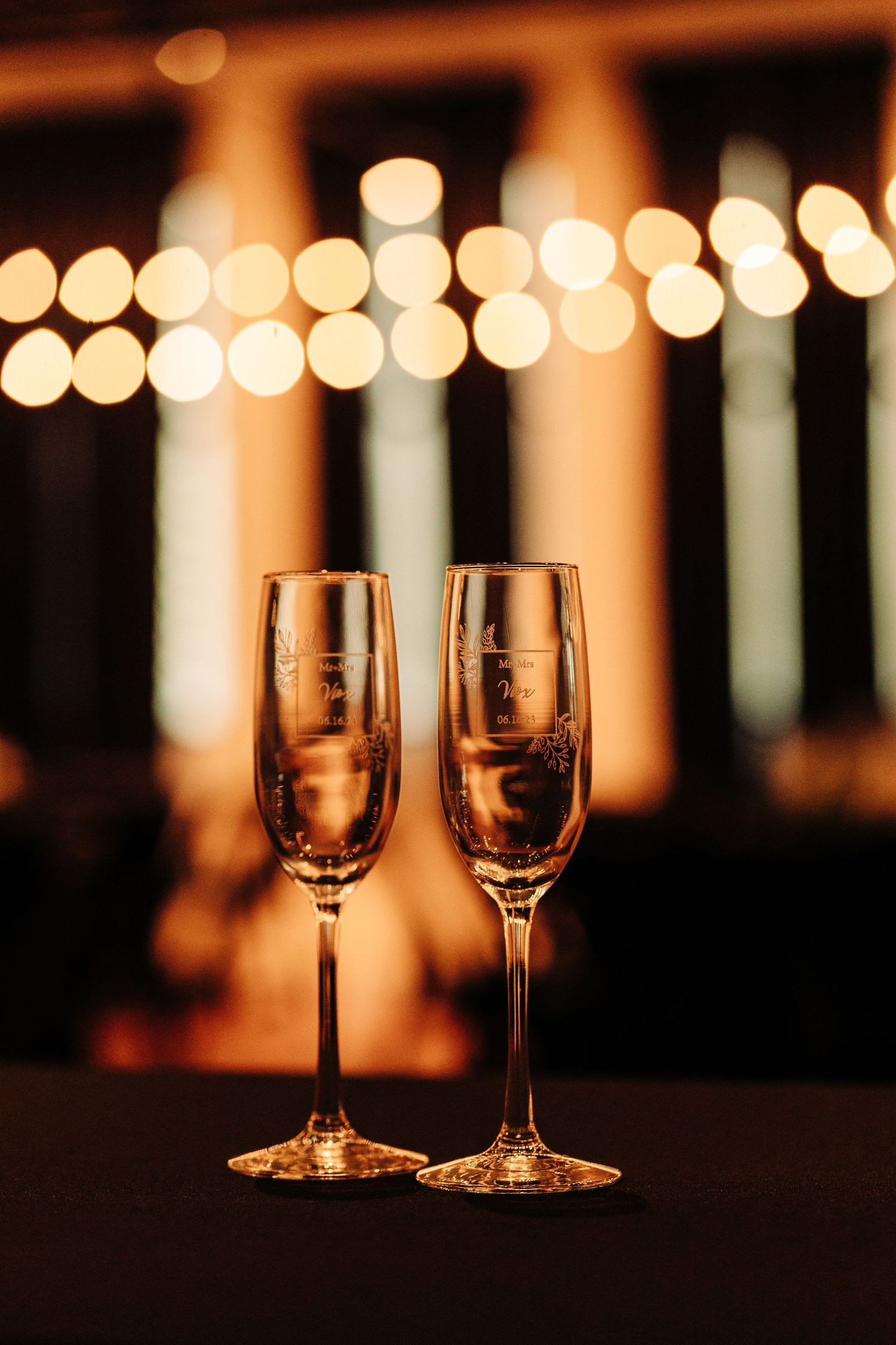 Two elegant wine glasses sit against a warm bokeh background with glowing string lights creating a romantic ambiance.