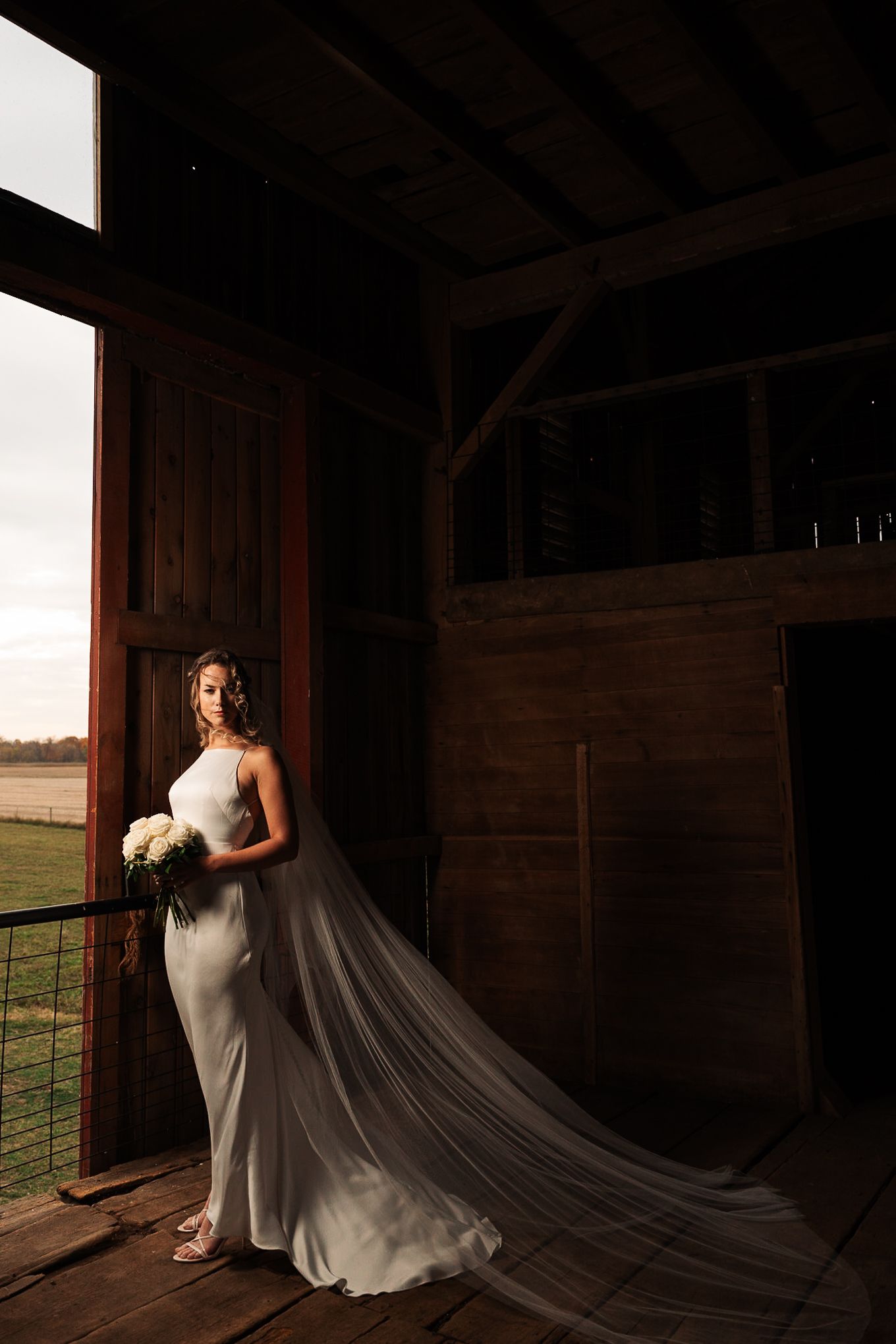A bride in a white gown with a flowing cathedral veil poses dramatically in a rustic barn doorway at sunset.