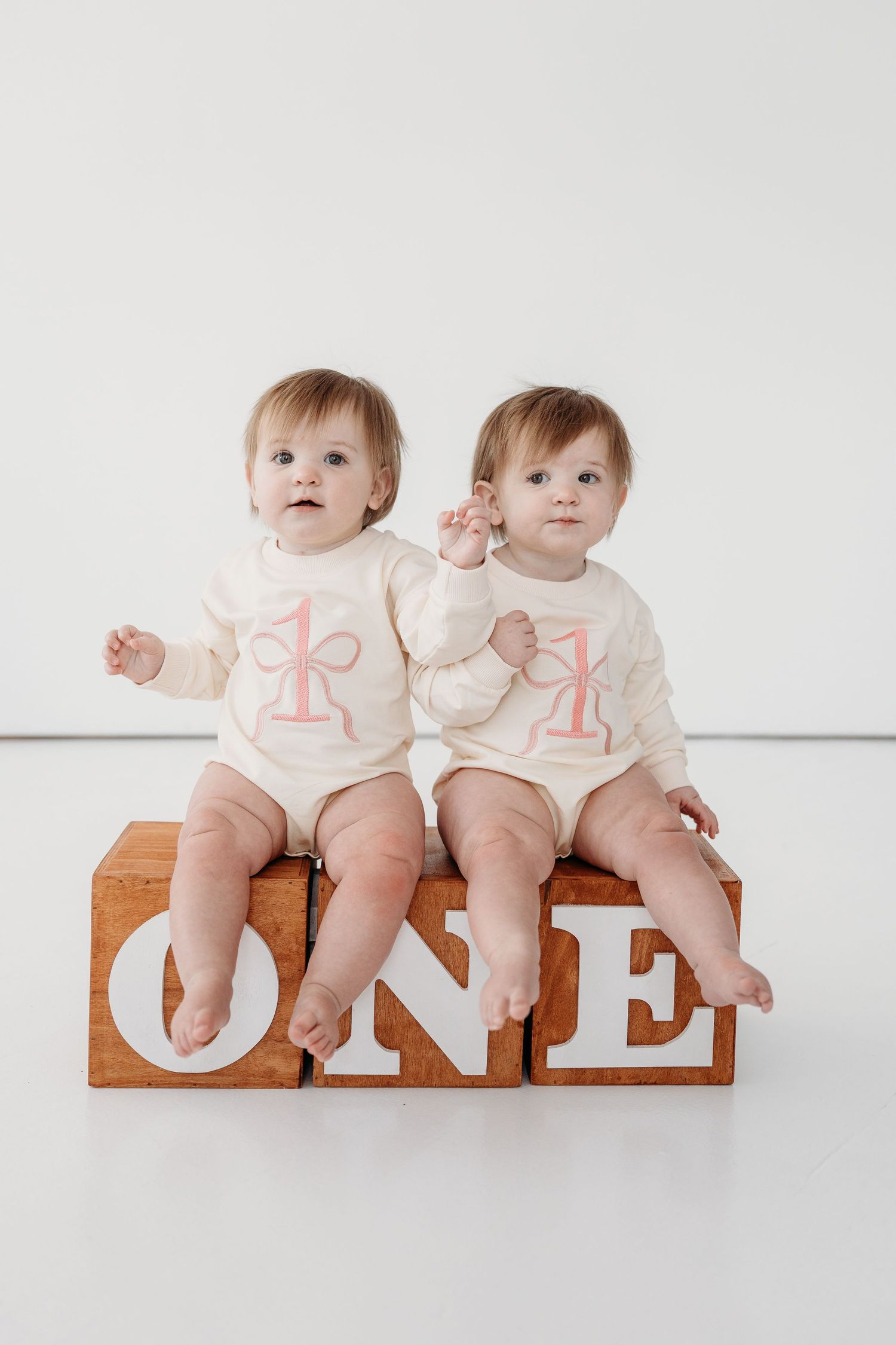 Two babies in matching white onesies with number one designs sit together on a wooden surface for their first birthday photo.