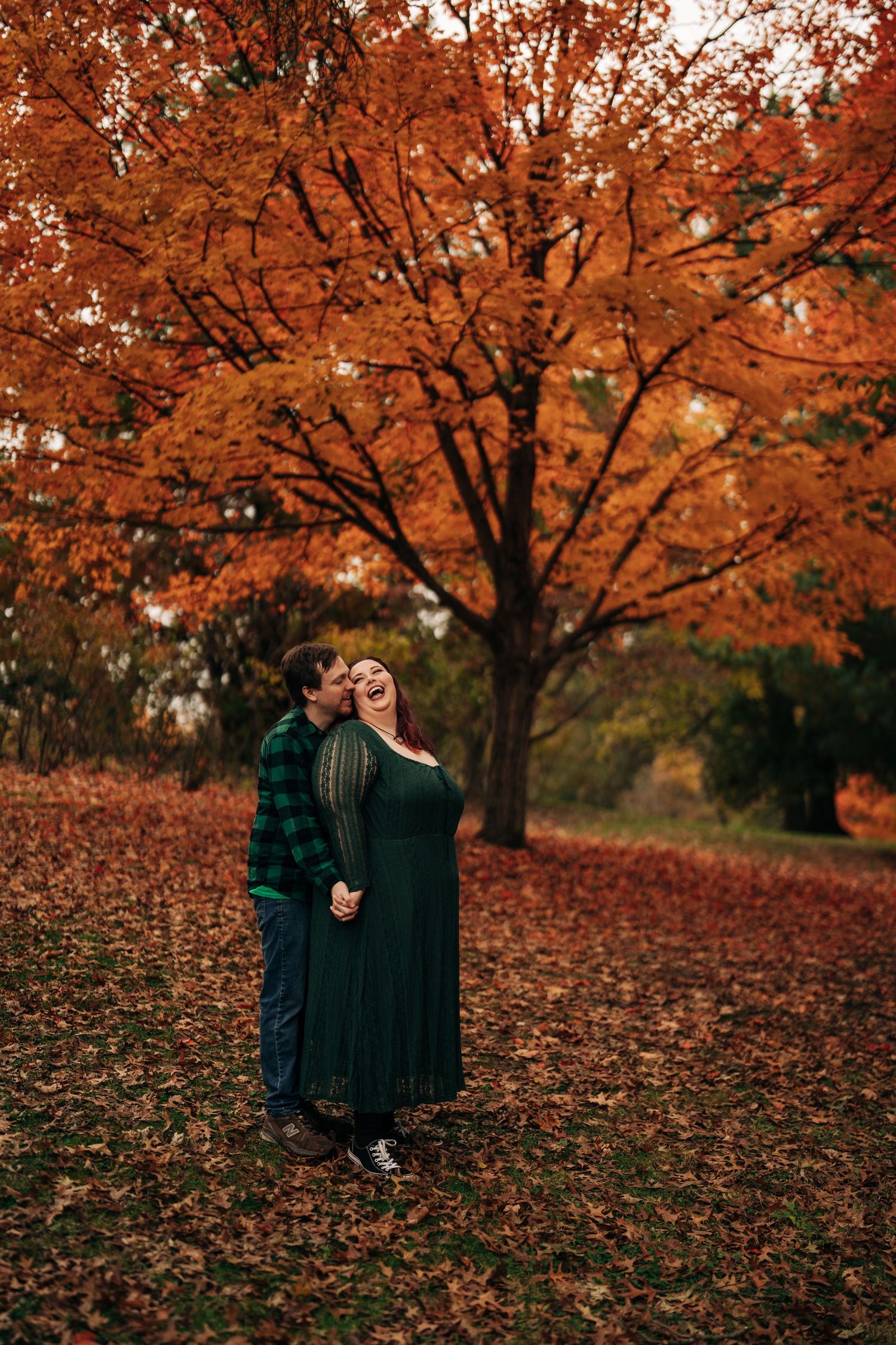 A couple embraces under a vibrant orange autumn tree in a park filled with fallen leaves.