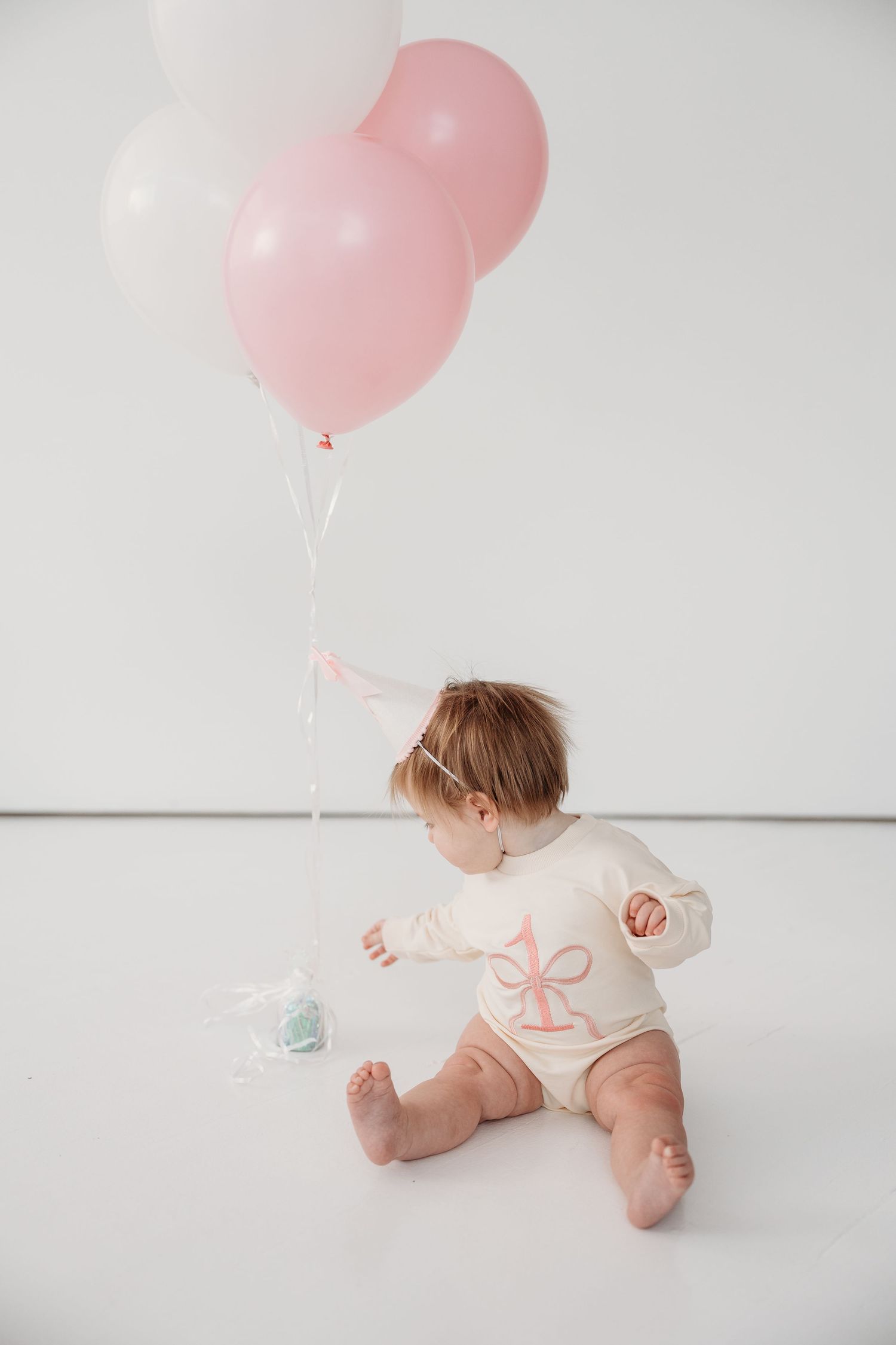 Baby in white outfit plays on floor with pink balloons against white background.