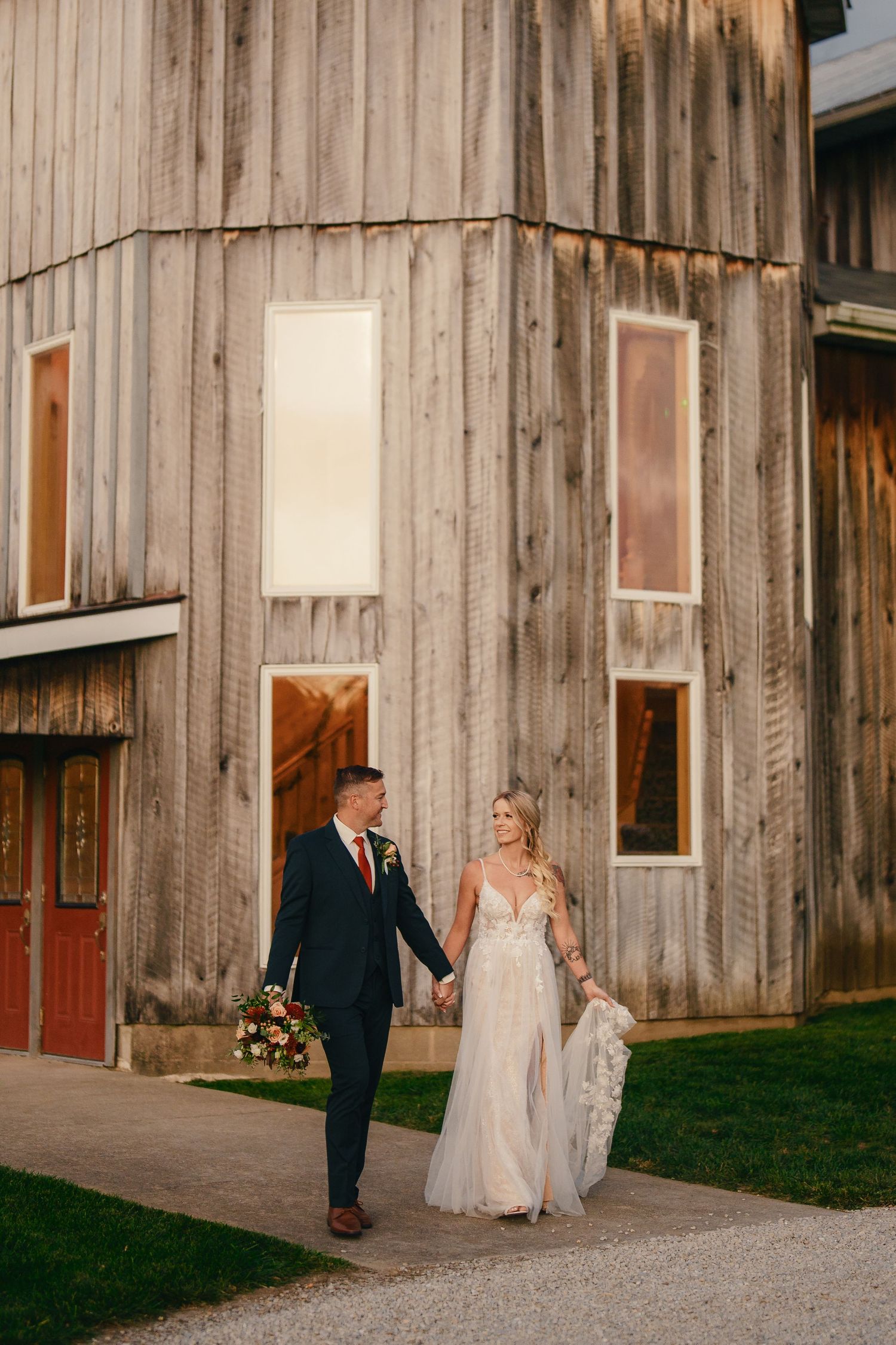 A couple in wedding attire walks together along the rustic exterior of a wooden barn venue.